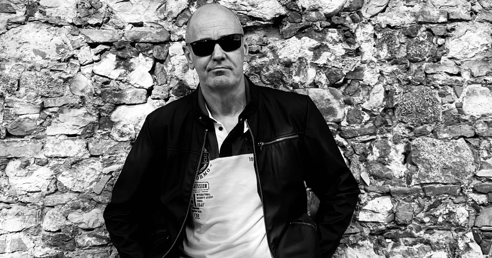 Schwarz-Weiß-Aufnahme des Künstlers Herbert Rutter, der vor einer Ziegelwand steht und eine Sonnenbrille trägt