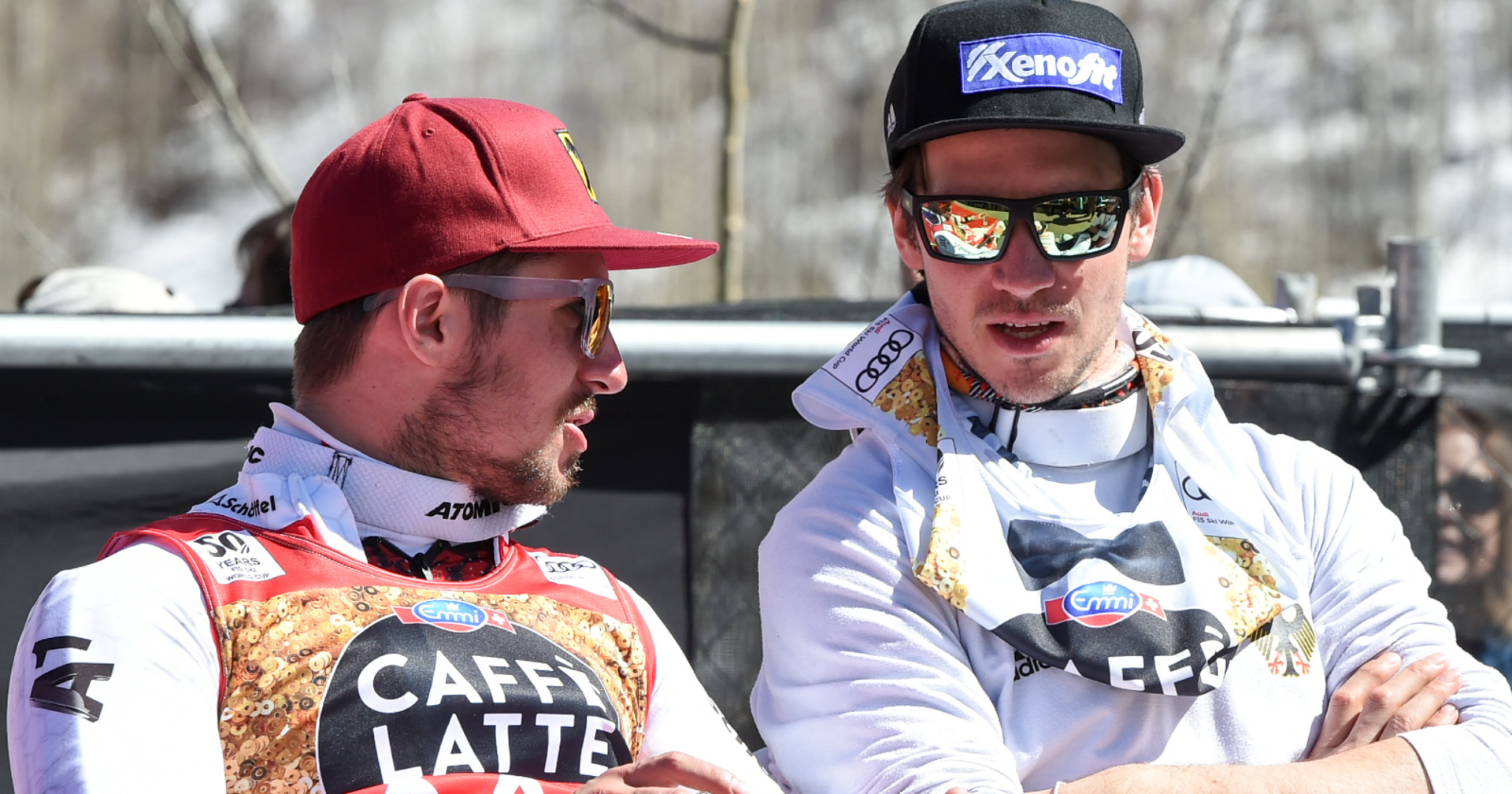 Marcel Hirscher und Felix Neureuther, die in Rennanzügen sowie mit Sonnenbrillen und Schirmmützen bekleidet, auf Stühlen sitzend miteinander diskutieren