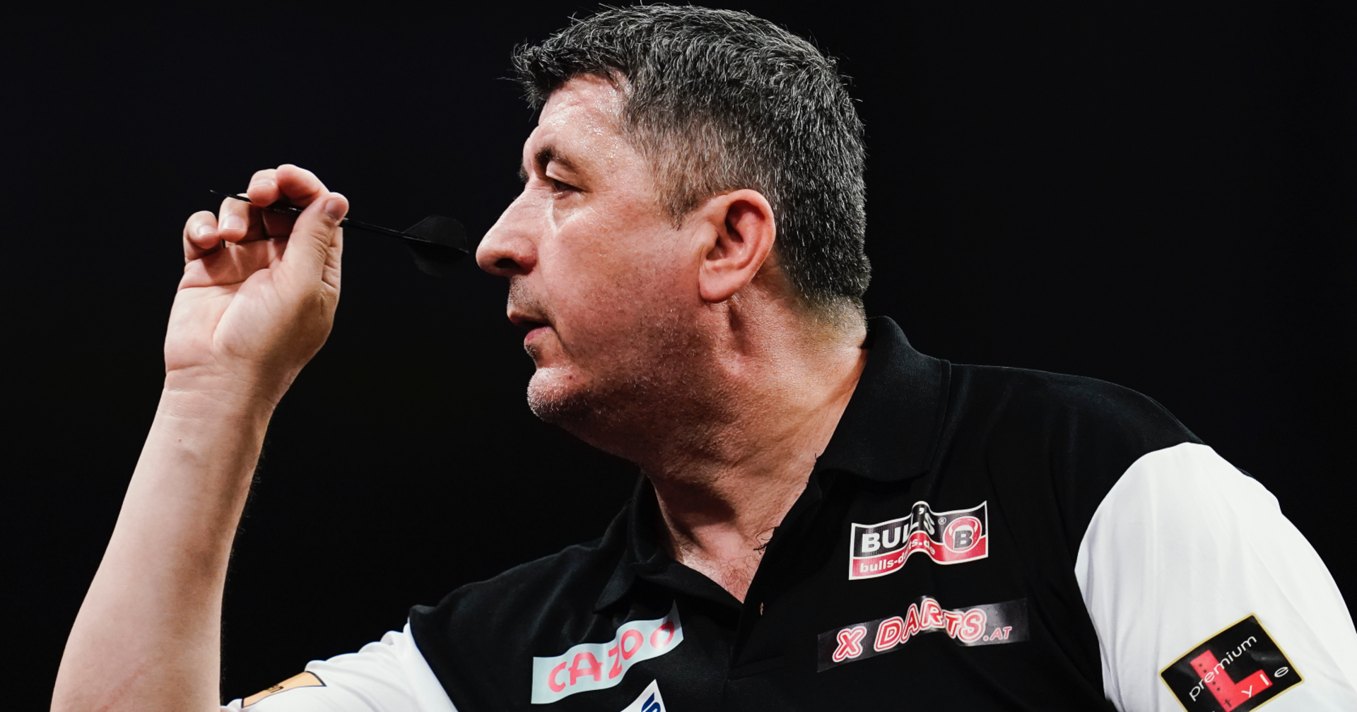 Nahaufnahme des Darts-Profis Mensur Suljović, der ein schwarzes Hemd mit weißen Ärmeln trägt und im Begriff ist, einen Dartspfeil zu werfen