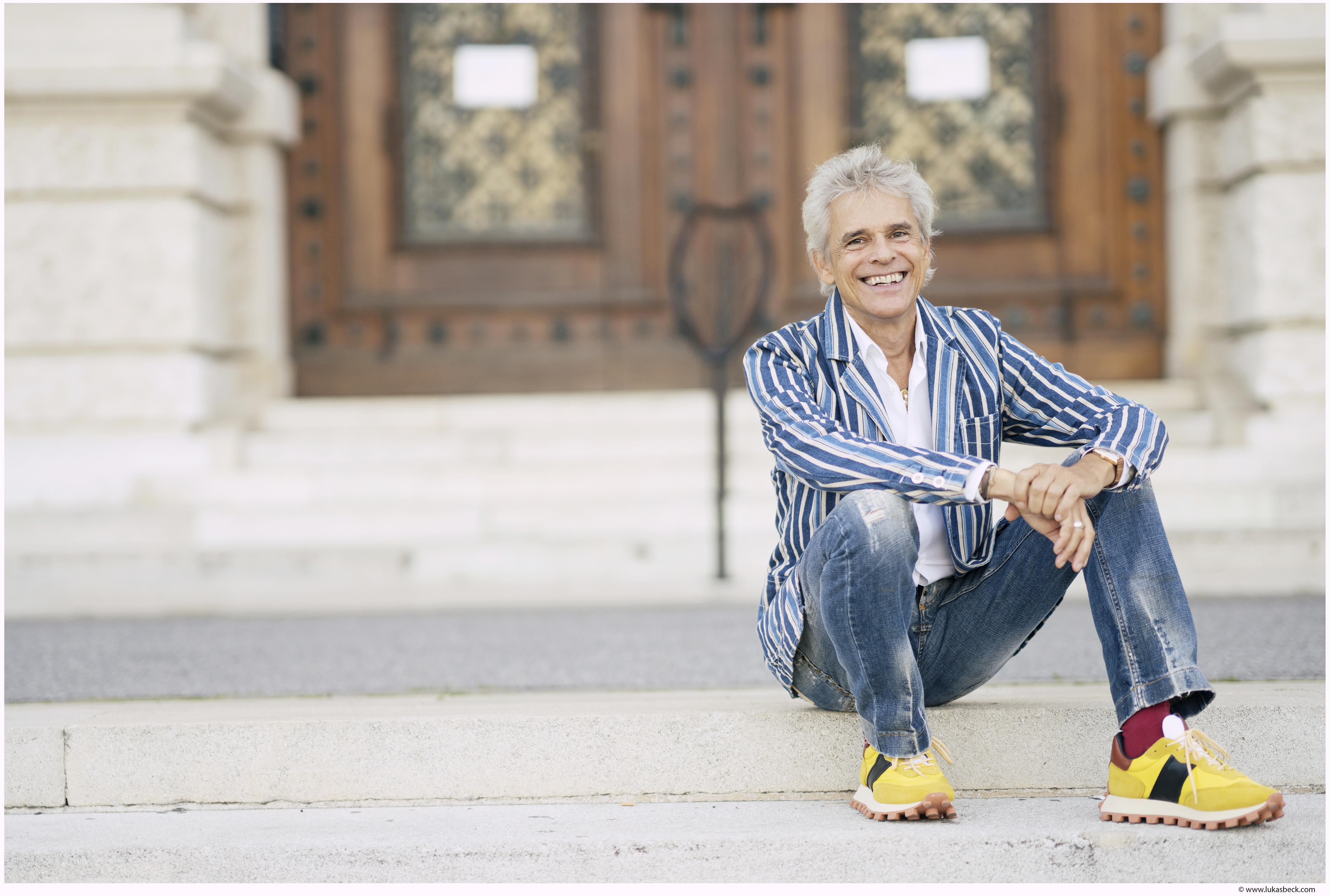 Thomas Brezina, der Jeans sowie ein blau-weiß-gestreiftes Sakko trägt, und auf Treppenstufen vor einem Hauseingang sitzt