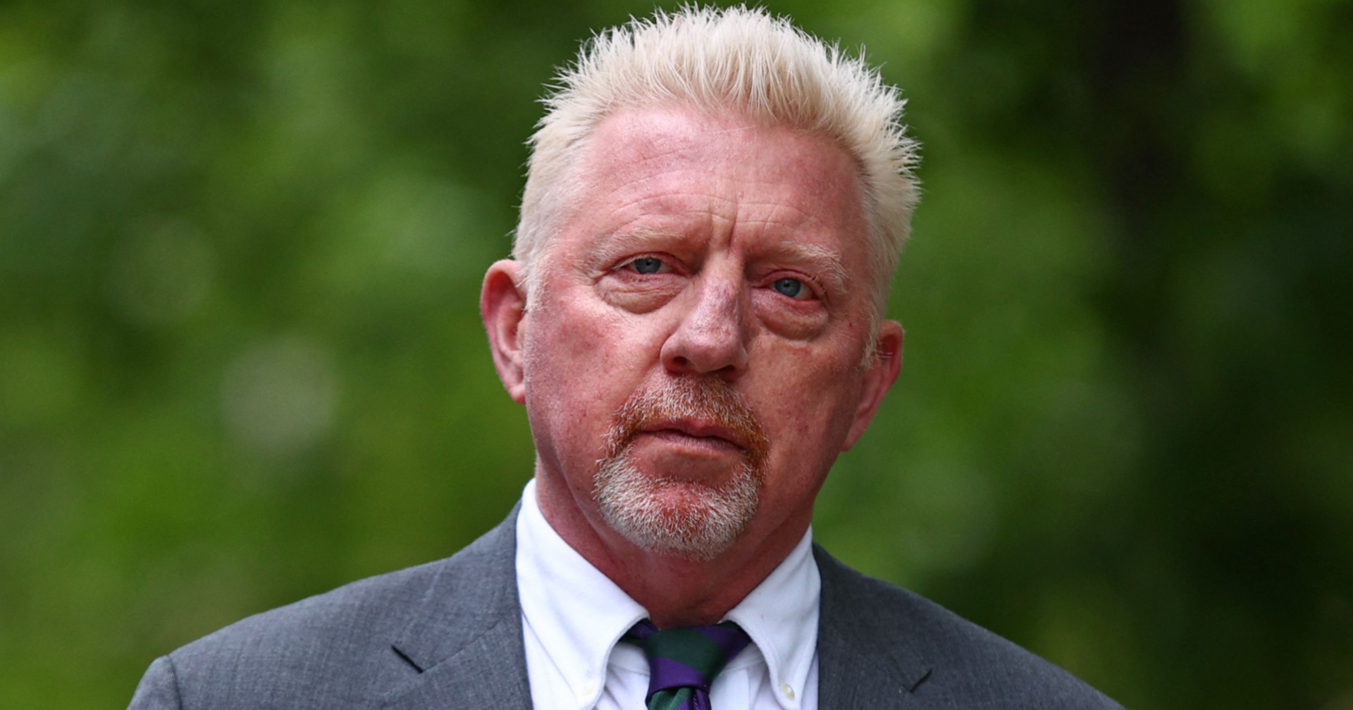 Porträt von Boris Becker, der einen grauen Anzug, ein weißes Hemd sowie eine grün-violett gestreifte Krawatte trägt