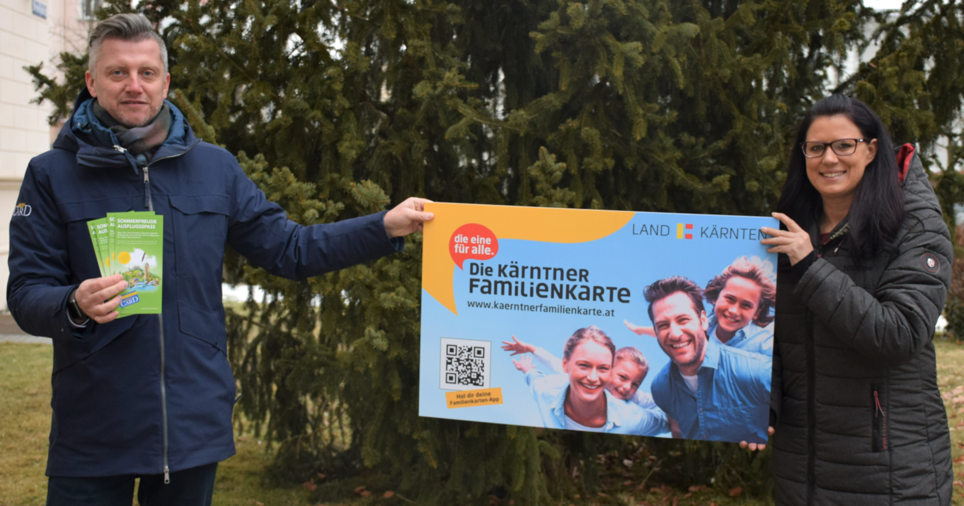 Sara Schaar und Anton Fasching halten vor einem Baum im Garten der Landesregierung das überdimensional große Modell einer Kärnten Card in die Höhe