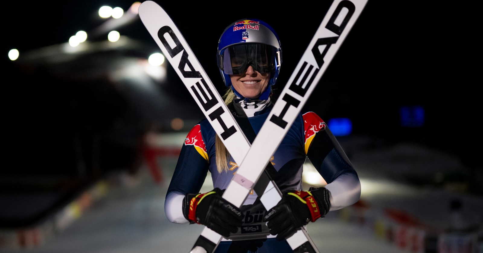 Lindsey Vonn im Zielhang der Streif bei Nacht. Sie trägt einen Rennanzug, einen Sturzhelm sowie eine Skibrille und hält zwei vor sich in den Schee gestellte weiße Head-Ski vor ihrem Oberkörper über Kreuz