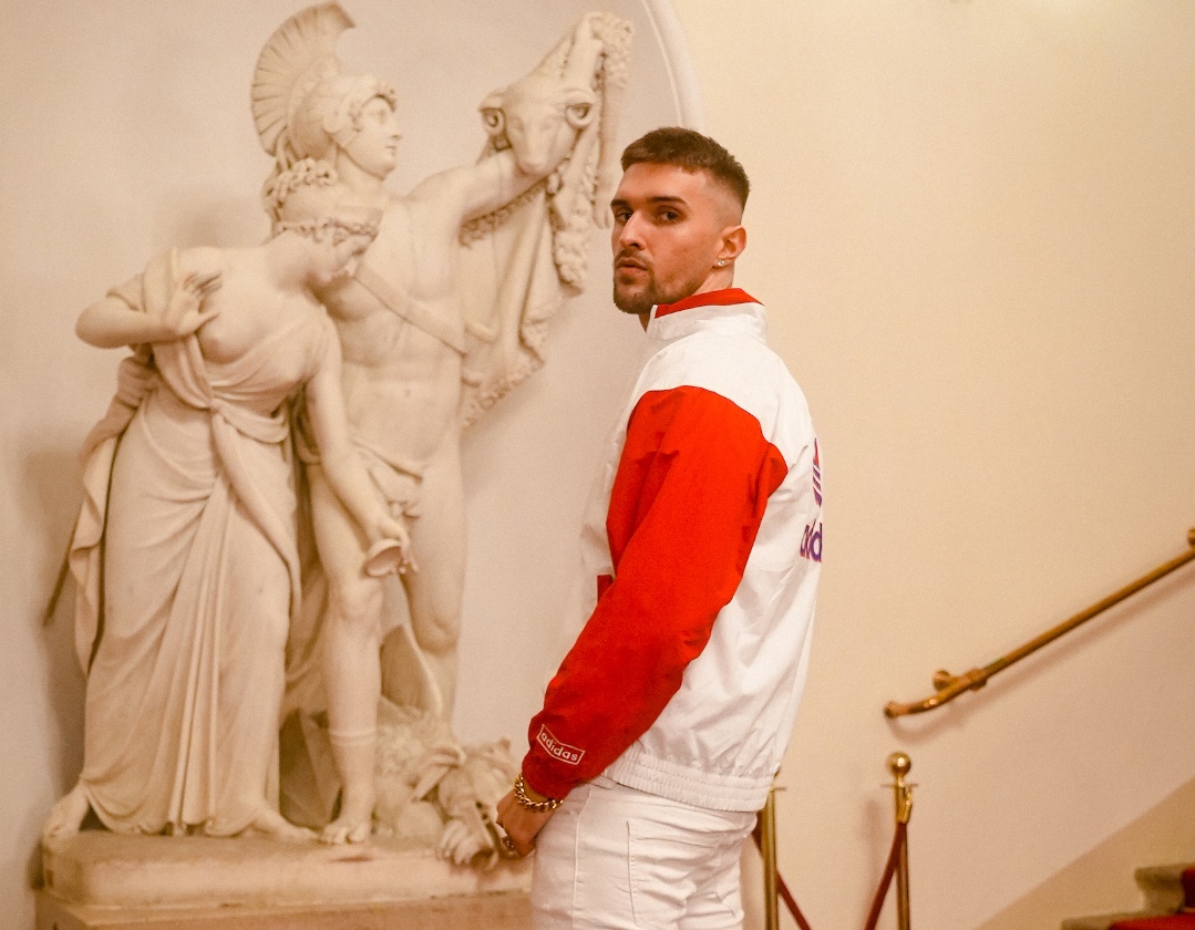 Der Rapper Gavis Dean, der in eine weiße Jacke mit roten Ärmeln gekleidet, vor einer Marmorstatue steht