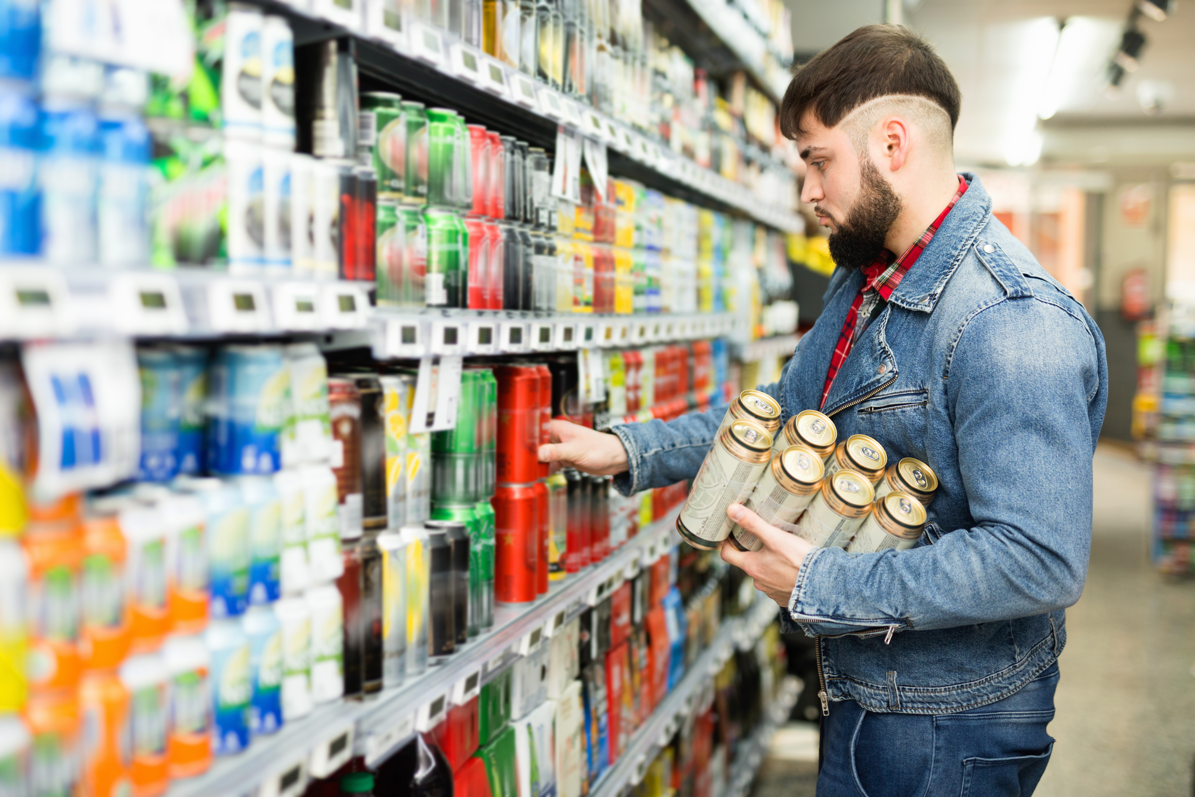 Ein junger Mann mit Vollbart und Jeansjacke entnimmt einem Supermarktregal acht Dosen Bier