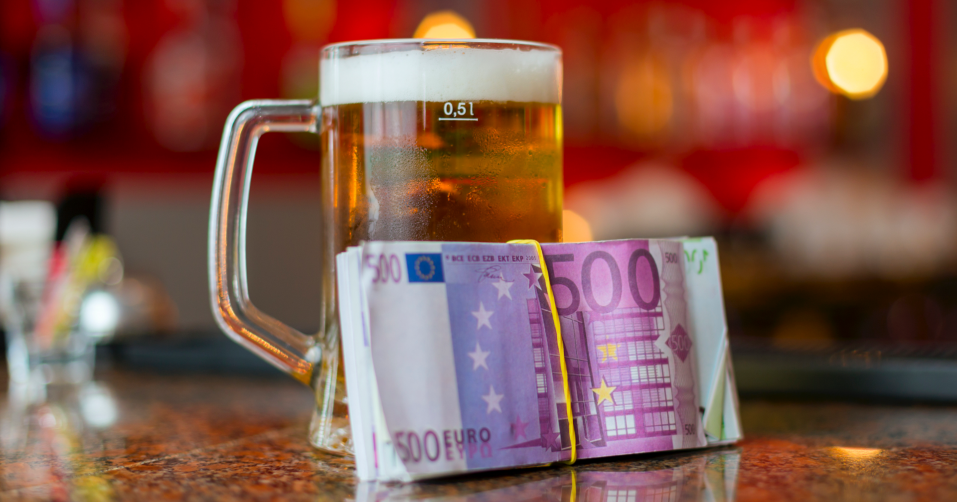 Ein Glas Bier auf einer Theke, an dem ein Bündel 500-Euro-Scheine lehnt