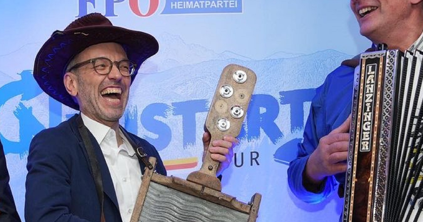 Herbert Kickl mit Hut spielt lachend auf einem Instrument vor der Pressewand der FPÖ. Neben ihm ein Mann mit Ziehharmonika