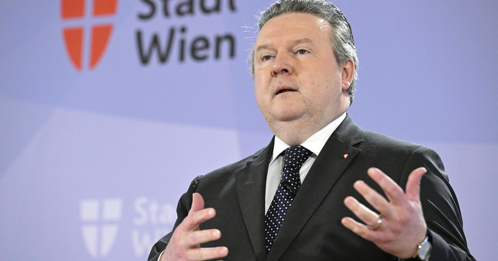Michael Ludwig im schwarzen Anzug mit Krawatte vor der Pressewand der Stadt Wien. Er spricht und gestikuliert