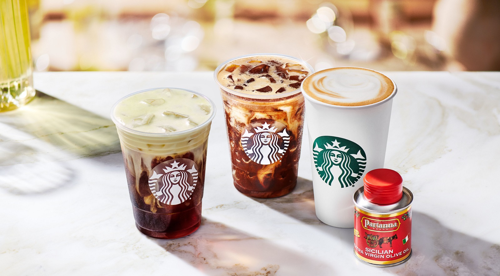 Drei verschiedene Starbucks-Getränke im entsprechenden Becher. Daneben eine kleine Flasche Olivenöl