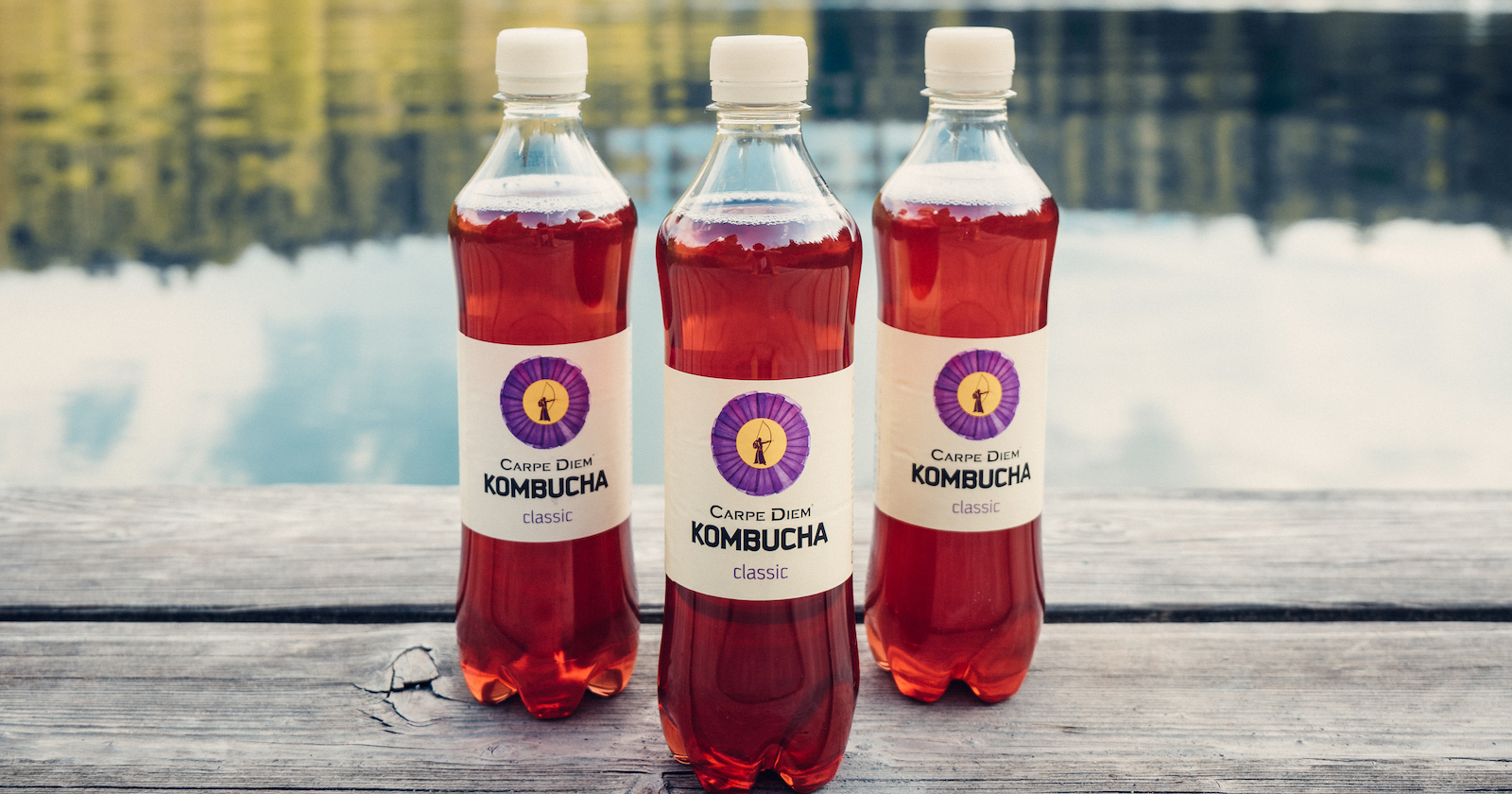 Drei Flaschen Kombucha vor einem See.