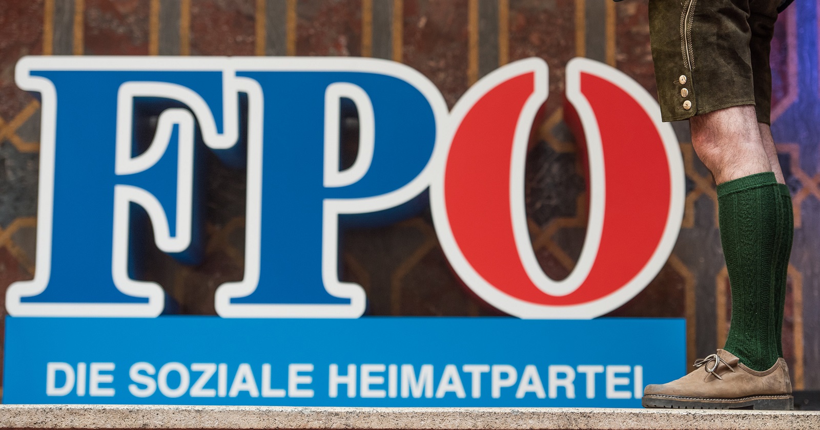 Das Logo der FPÖ mit dem Claim &quot;Die soziale Heimarpartei&quot; als Buchtaben auf einer Bühne stehen. Danebeb in einer Lederhose und Stutzen bekleidete Männerbeine.
