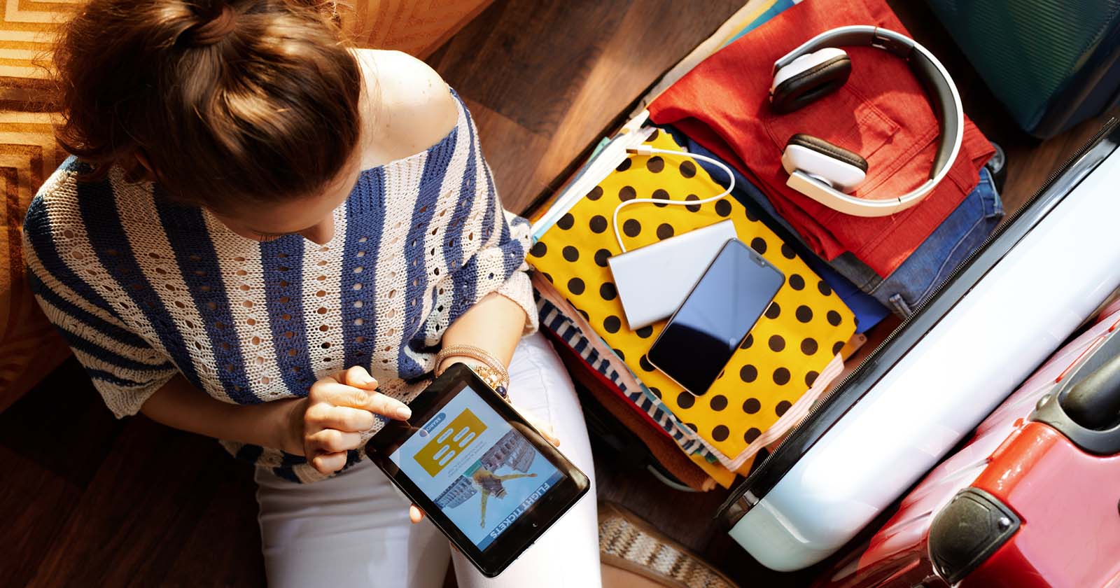 Frau sitzt vor einem gepackten Koffer und bucht auf dem Tablet eine Reise