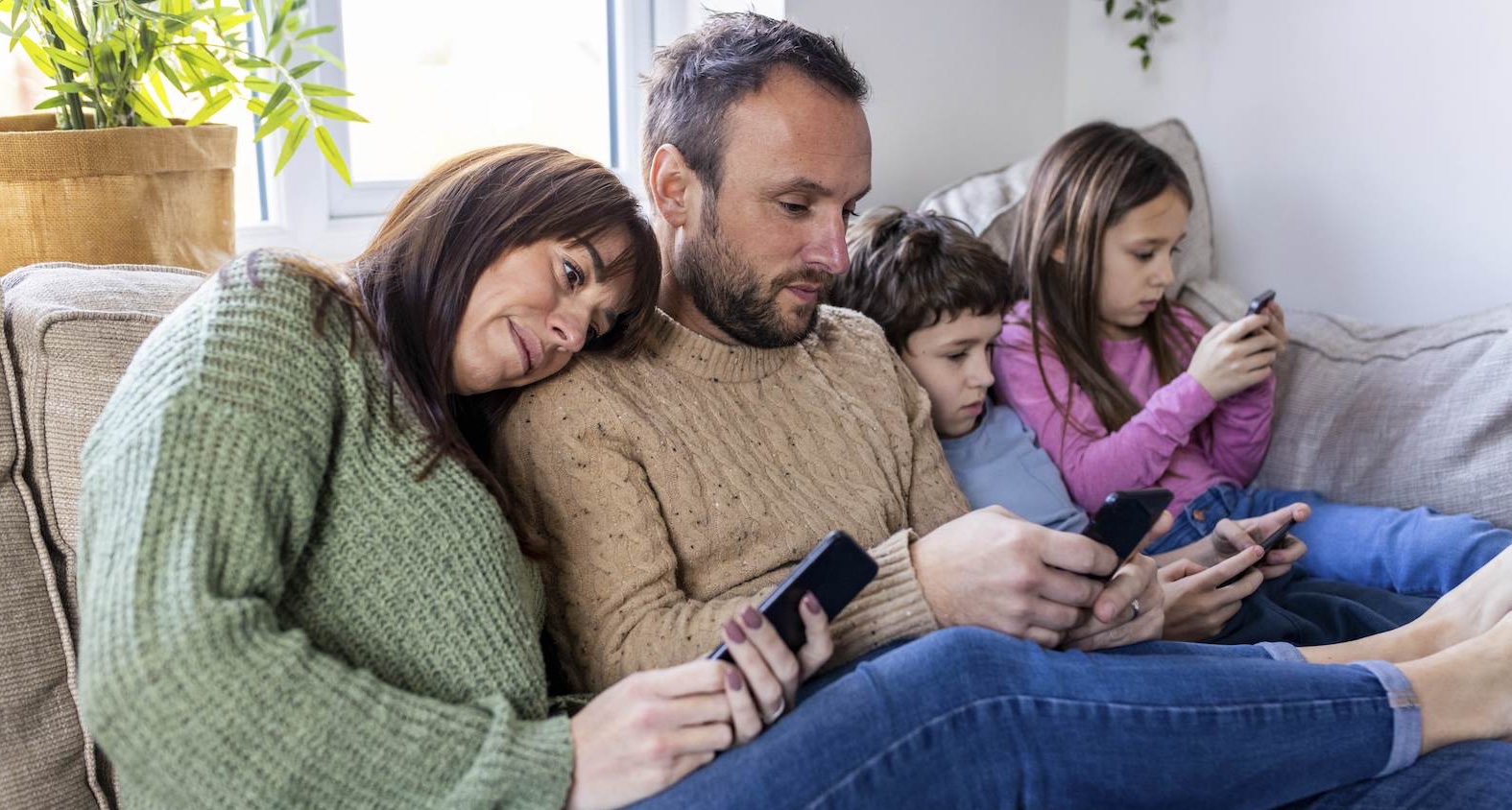 Mutter, Vater und zwei Kinder sitzen auf dem Sofa und schauen jeder in ein Smartphone
