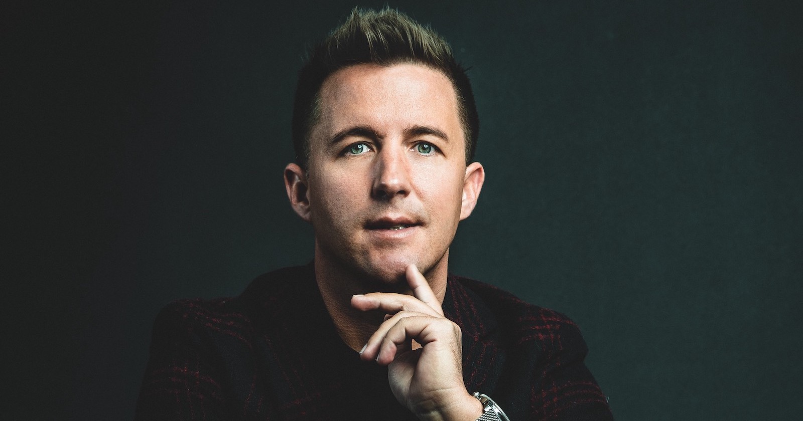 Porträt von Thomas Semmler vor einem schwarzen Hintergrund, der mit seinem Zeigefinger gegen sein Kinn tippt