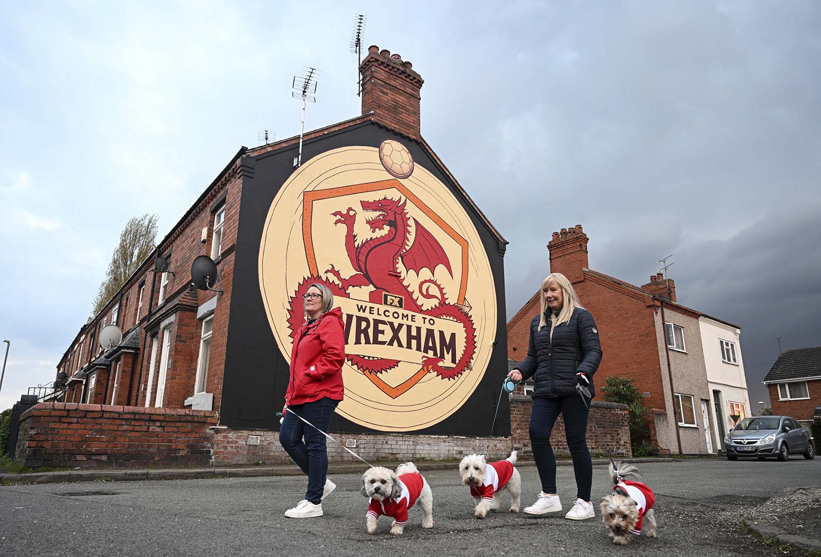 Zwei Frauen mit drei Hunden gehen an einem Haus mit einem Logo des AFC Wrexham vorbei.