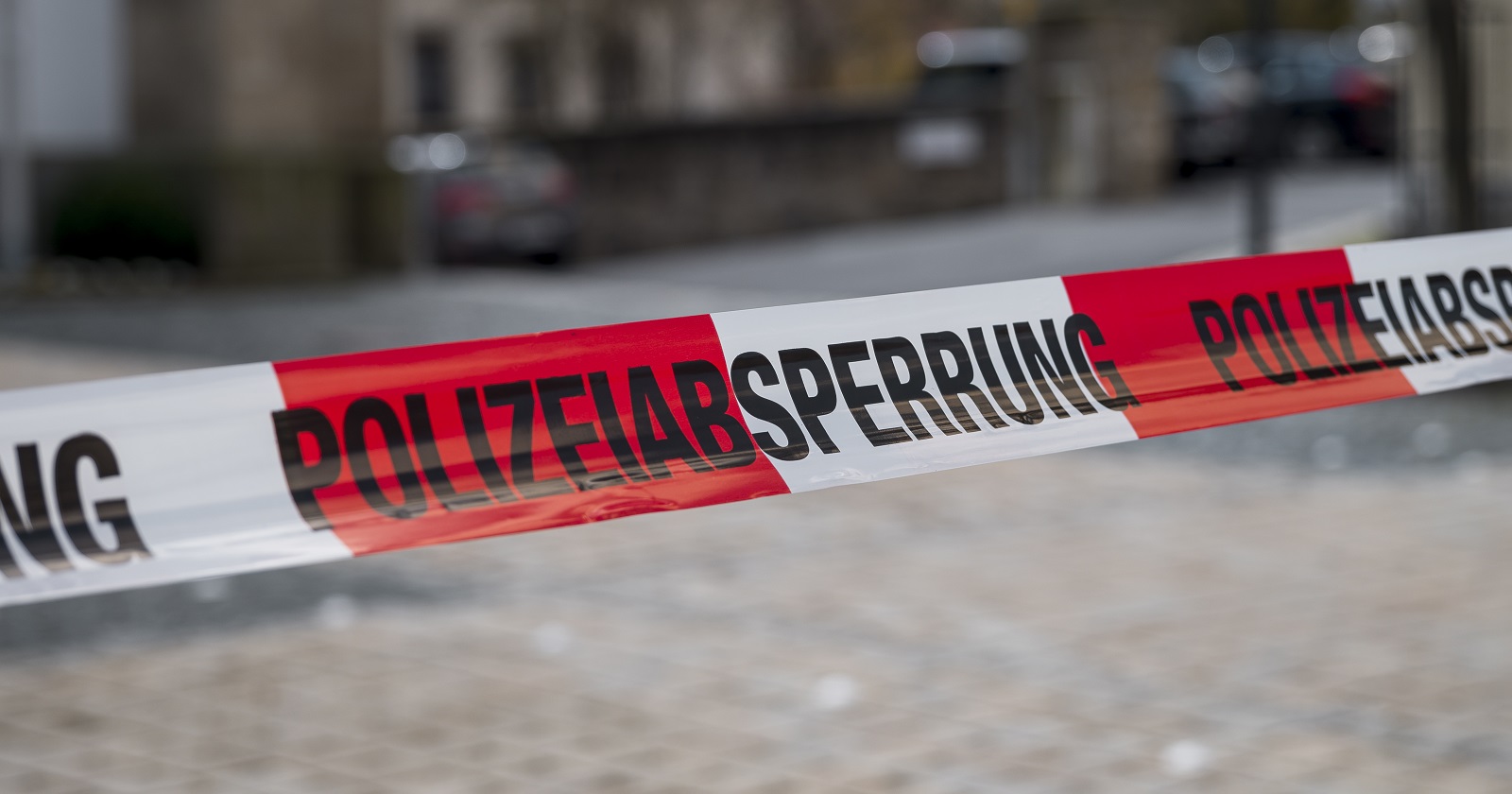 Ein rot-weiß-rotes Absperrband mit dem Schriftzug &quot;Polizeiabsperrung&quot; auf eine Straße mit Kopfsteinpflaster.
