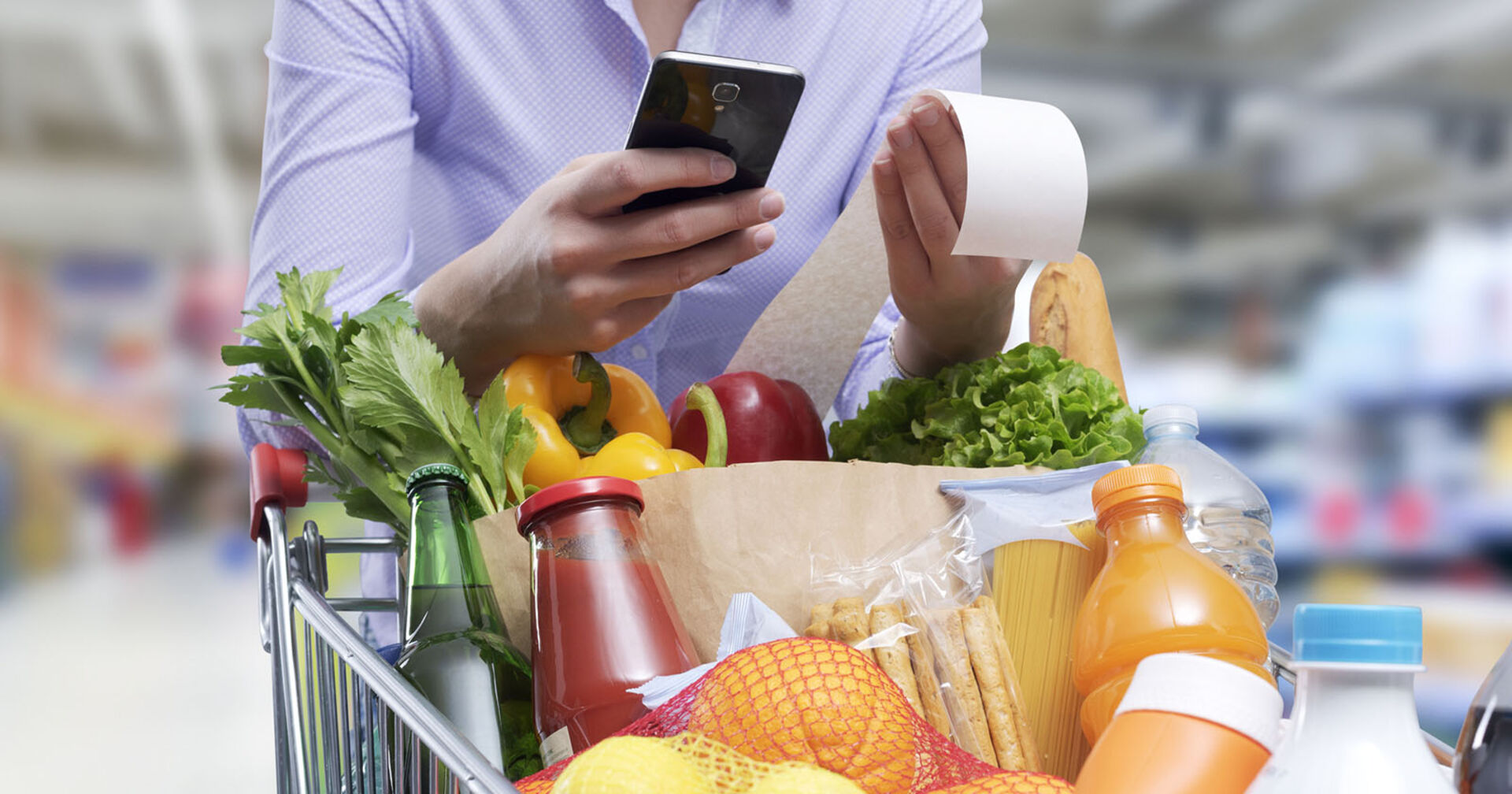 Frau mit einem gefüllten Einkaufswagen prüft mit ihrem Smartphone die Rechnung.