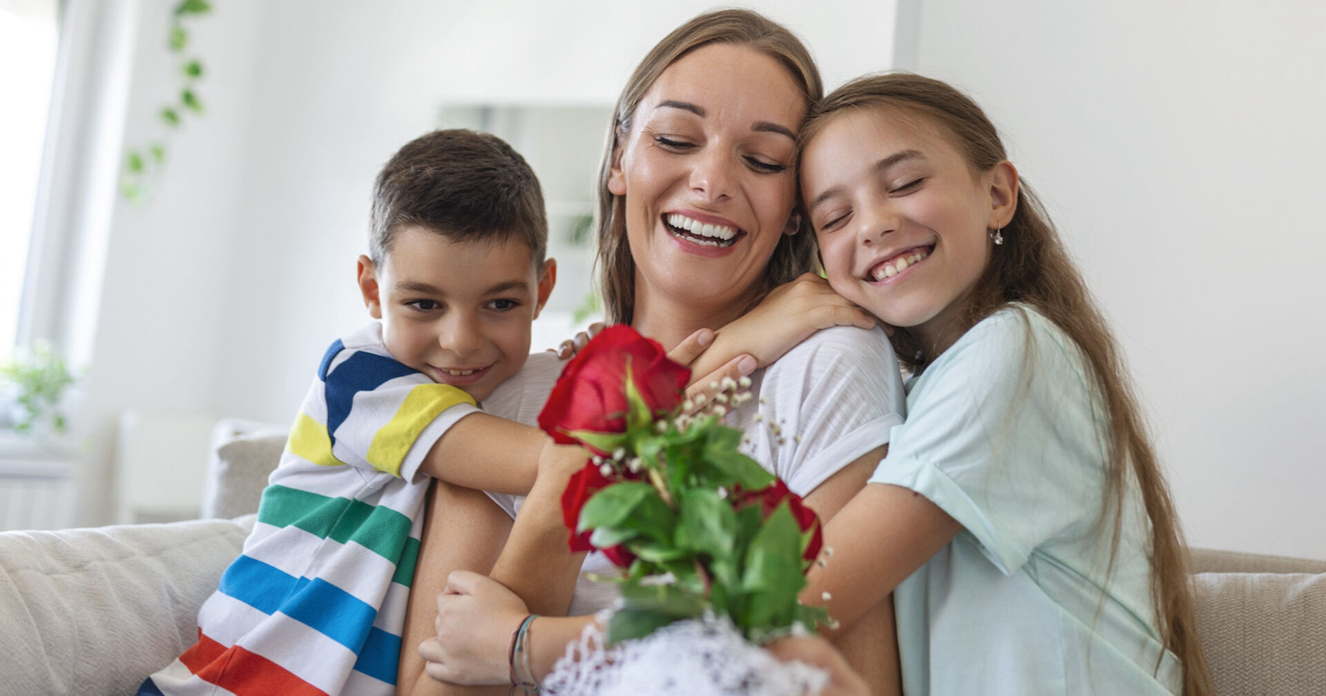 Sohn und Tochter umarmen ihre Mutter, die rote Rosen hält | Credit: istock.com/stefanamer