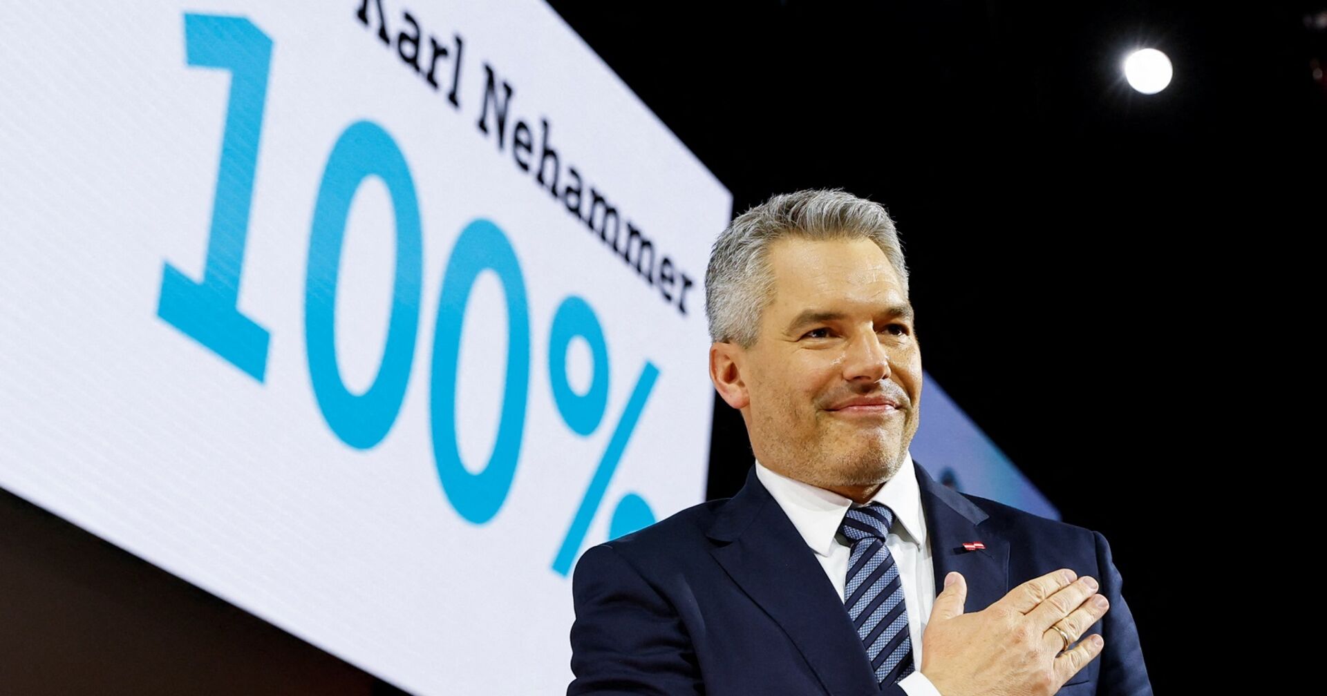 Karl Nehammer vor einem Schild mit der Aufschritt &quot;Karl Nehammer 100 Prozent&quot;. Er grinst und hat die Hand am Herz.