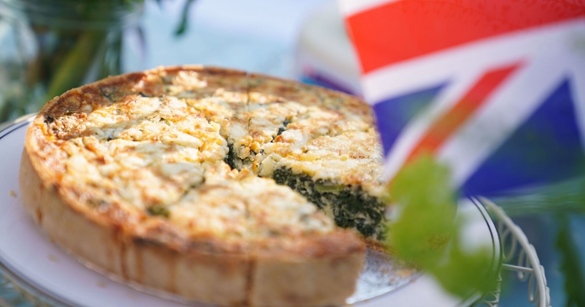 Eine angeschnittene Coronation Quiche, man sieht die Füllung. Im Vordergrund die britische Flagge.