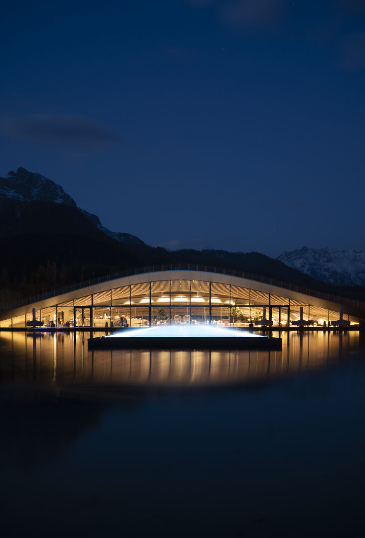 Beleuchtete Infinitypool im See, dahinter das beleuchtete Spa-Gebäude