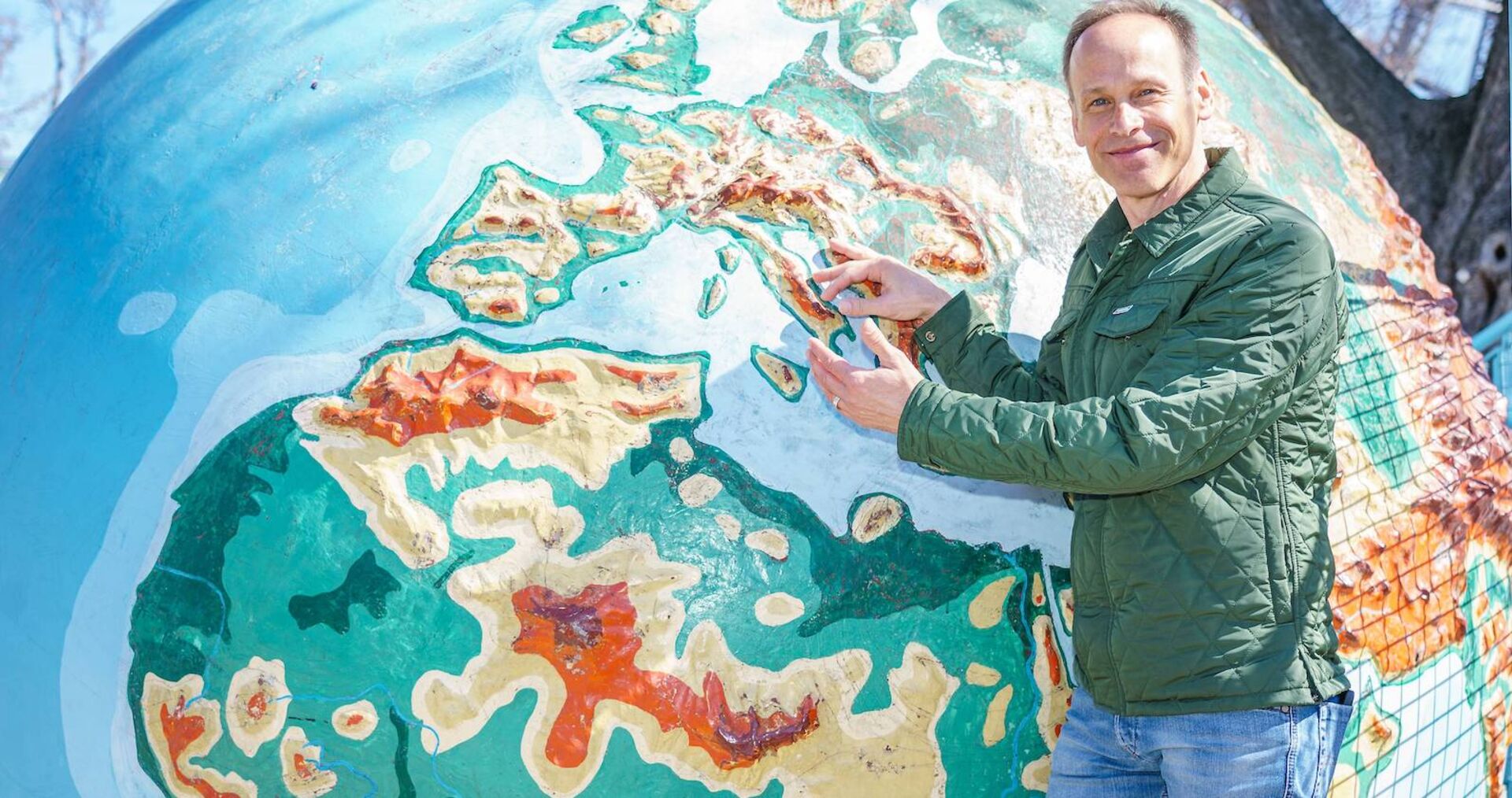 Meteorologe Marcus Wadsak steht vor einer Weltkugel und zeigt mit dem Finger auf ein Land