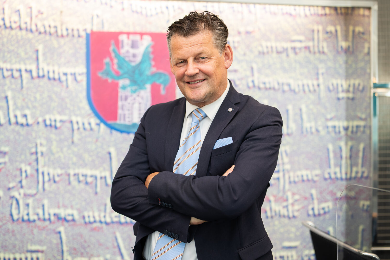 Bürgermeister Scheider, in Anzung und mit einer Krawatte, der mit verschränkten Armen vor dem Klagenfurter Stadtwappen steht