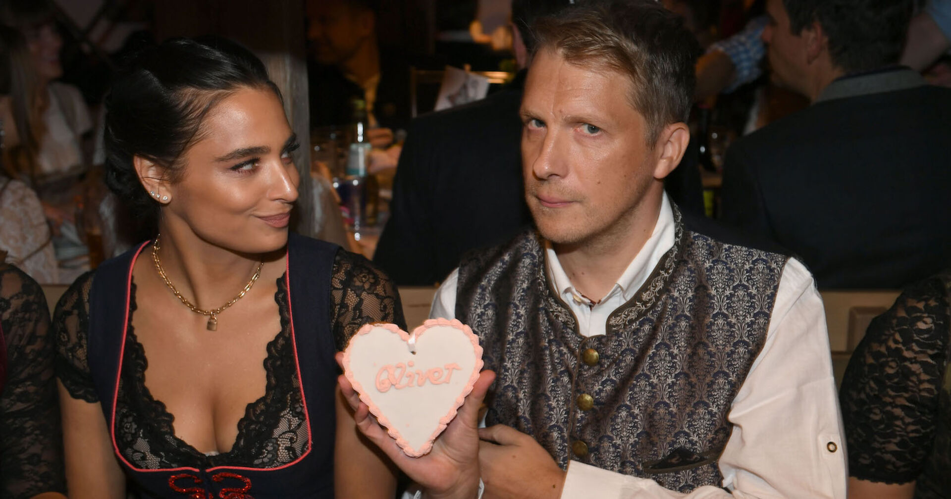Ein Paar sitzt am Oktoberfest, der Mann hält ein Herz in die Kamera.