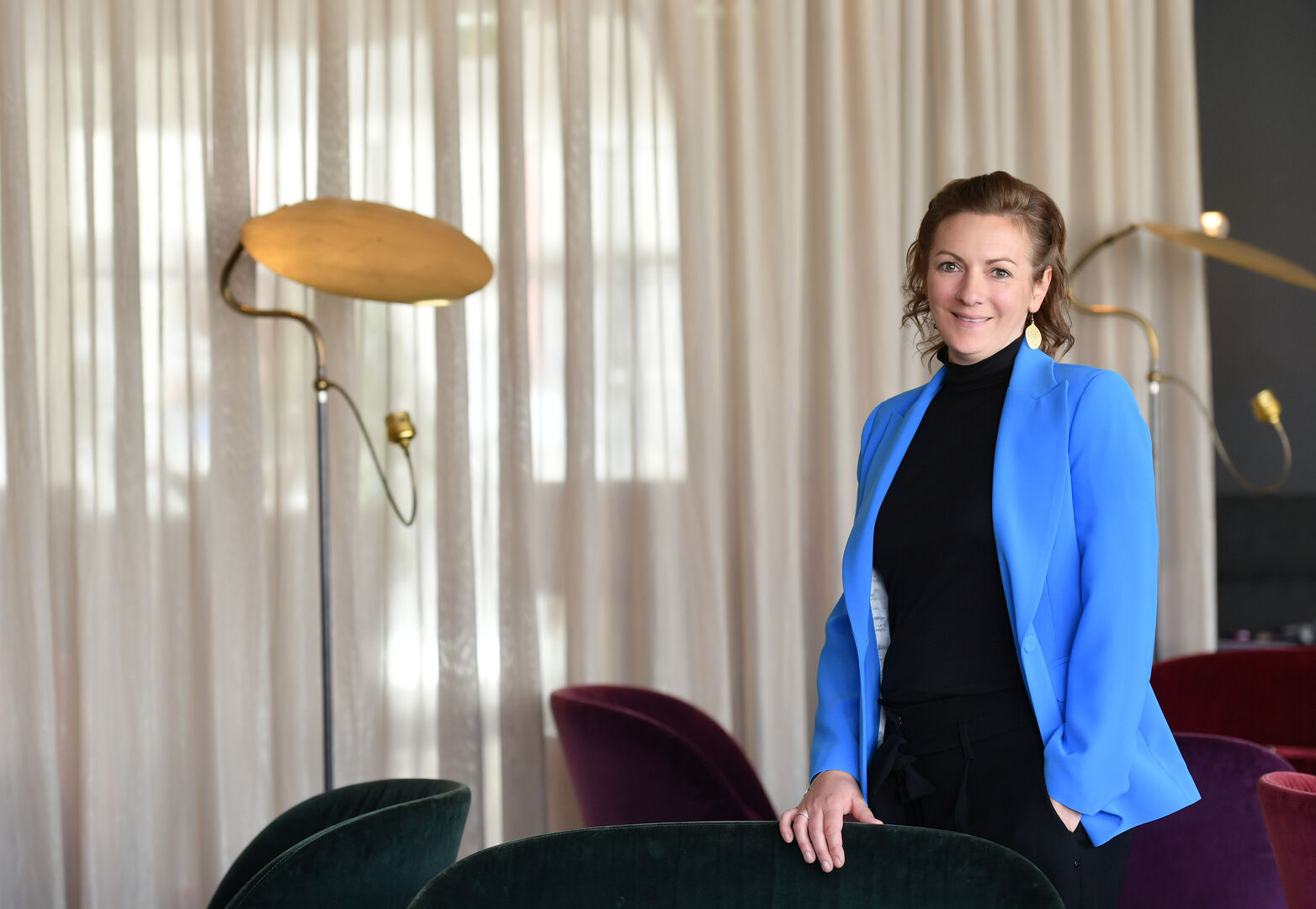 Karina Winkler, die ein schwarzes Business-Outfit samt blauem Sakko trägt, und zwischen mehreren Lounge-Stühlen steht