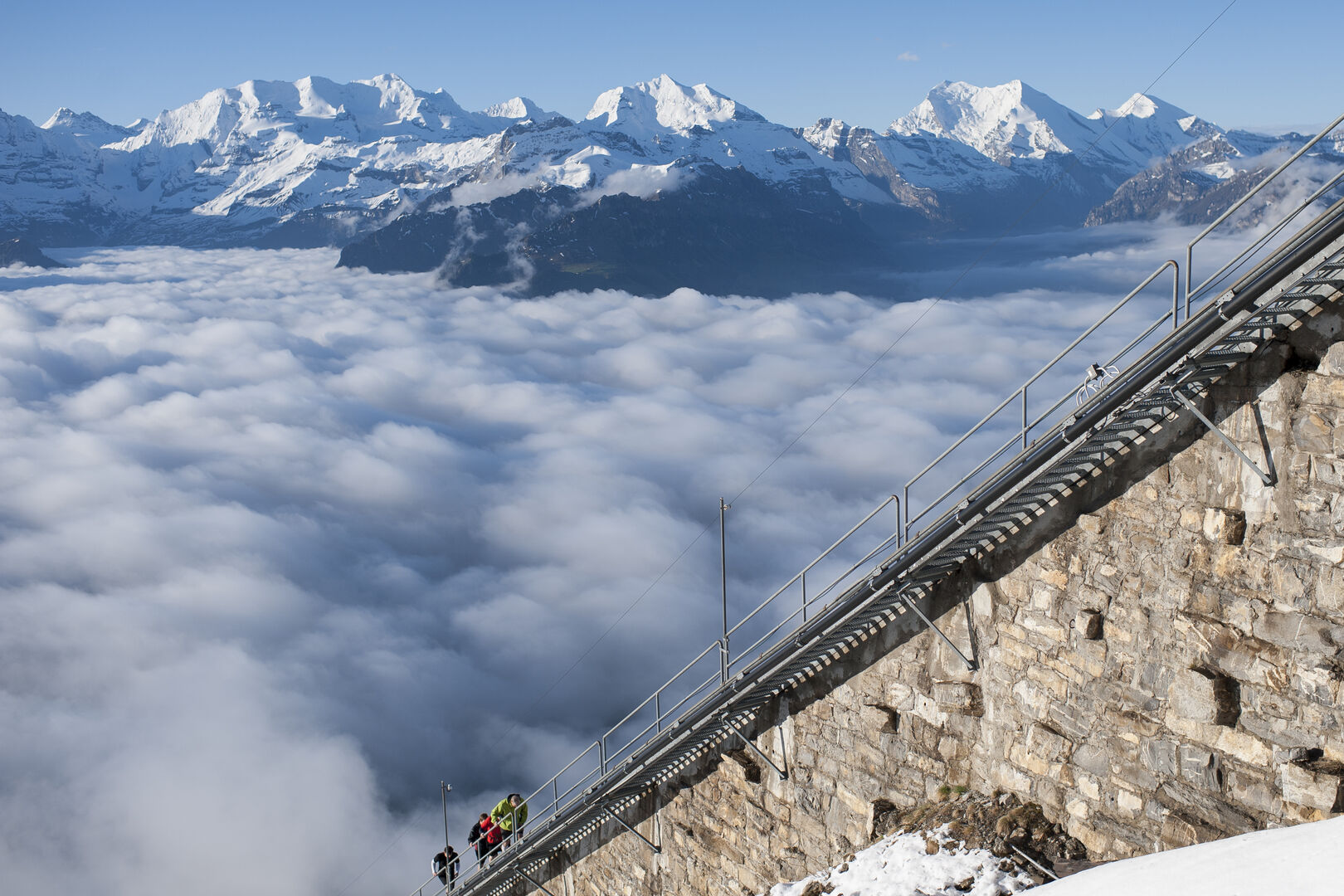 Bergwelt, Nebel und vorne im Bild drei Menschen, die eine steile Treppe erklimmen