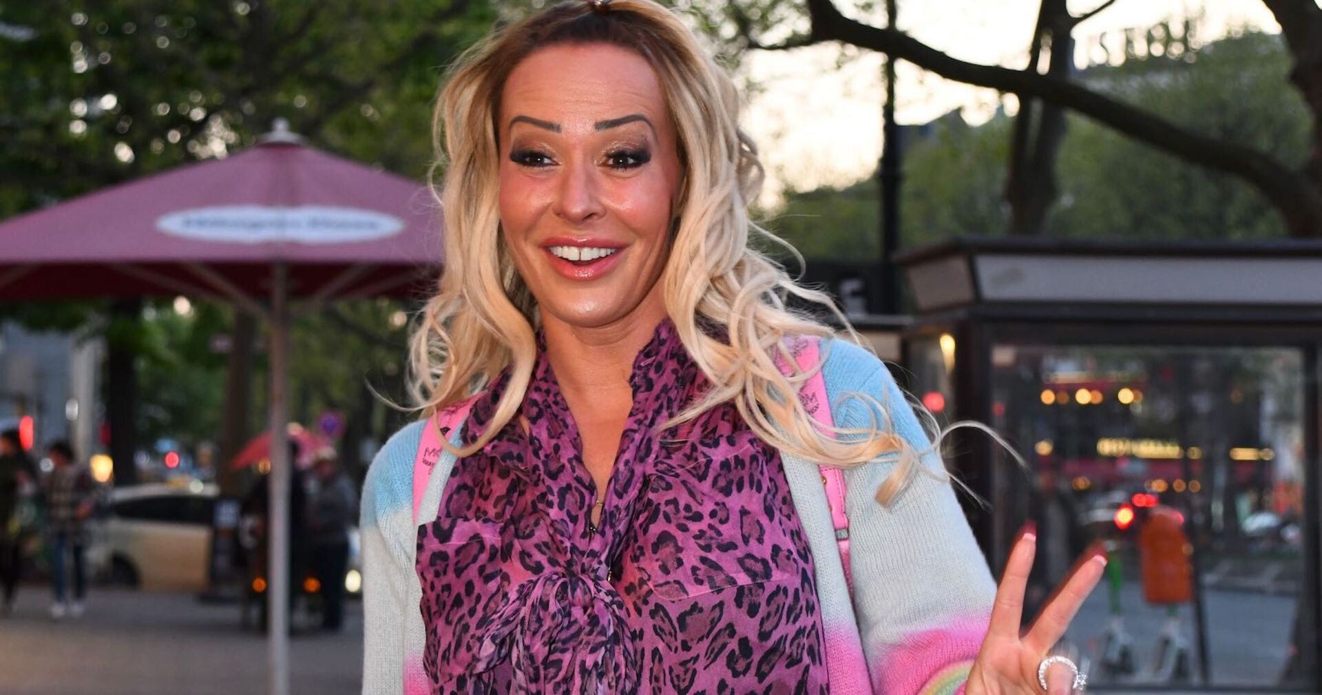 Eine blonde Frau mit einer rosa Leopardenbluse lacht in die Kamera.