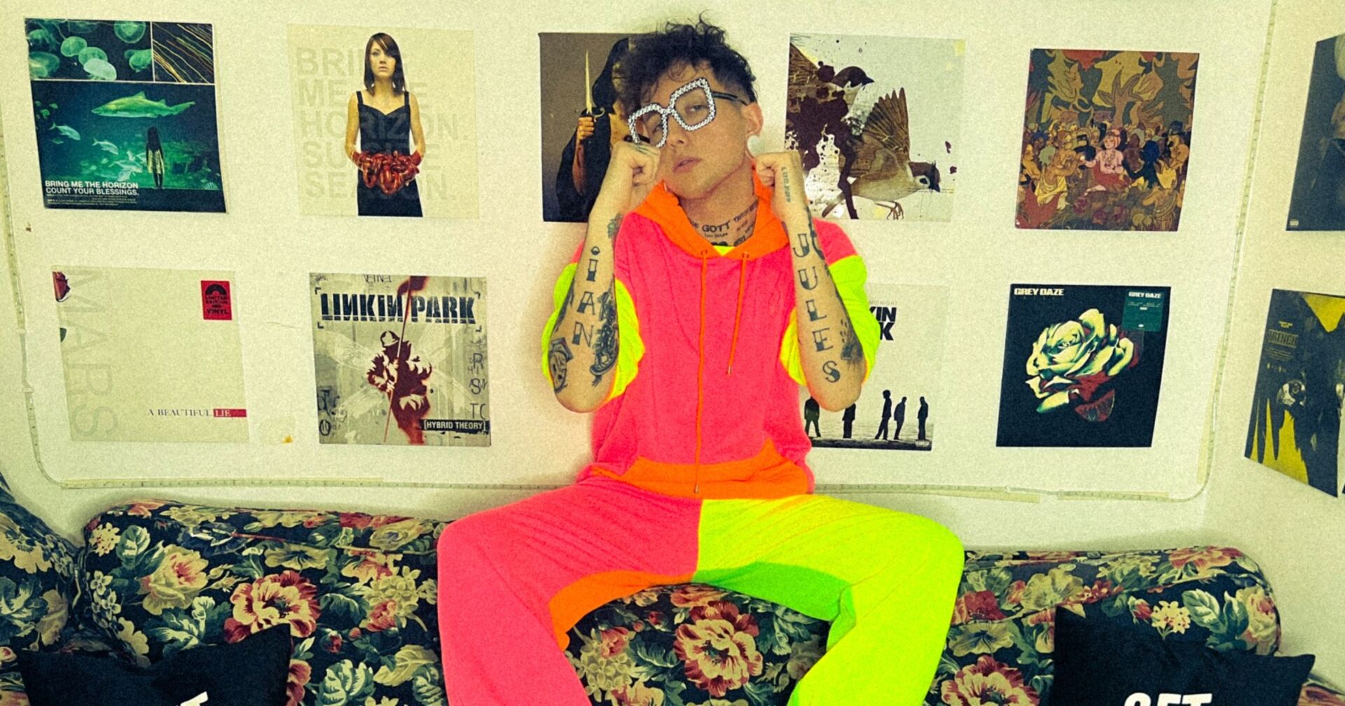 Ian Jules, der in einem neonfarbenen Outfit auf der Rückenlehne einer Couch sitzt. Im Hintergrund sind zahlreiche Plattencover an der Wand zu sehen.
