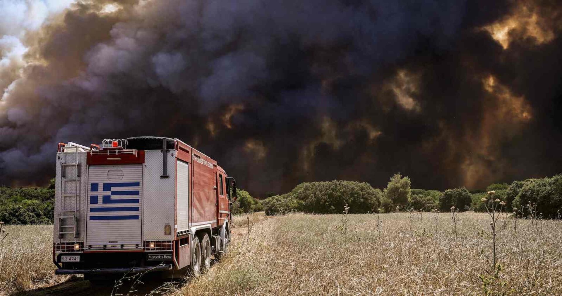 Ein Feuerwehrauto fähr über eine völlig vertrocknete Wiese auf eine Schwarze Wand aus Rauch und Flammen zu.