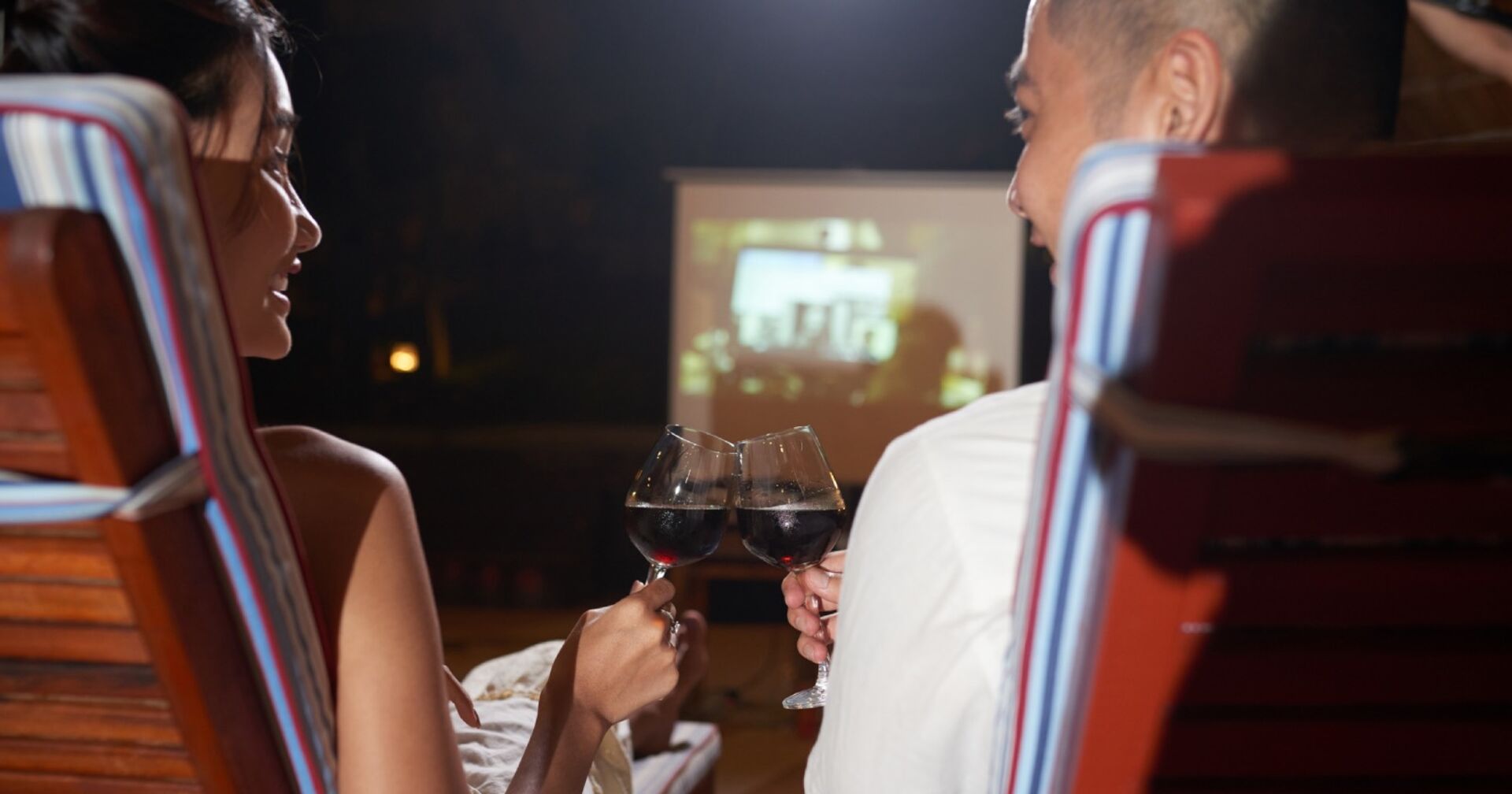 Ein Mann und eine Frau stoßen, in Stühlen sitzend, vor einer Kinoleinwand mit zwei Rotweingläsern an
