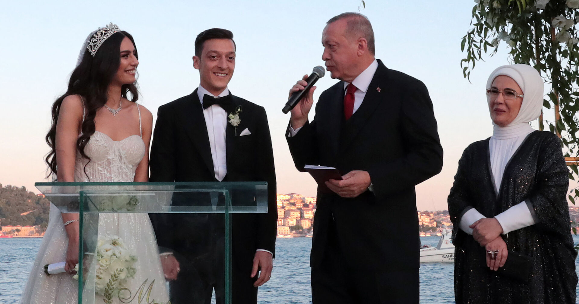 Özil und seine Frau am Traualtar, Erdogan steht neben ihnen und spricht ins Mikrofon, seine Frau an seiner Seite. Im Hintergrund das Meer.