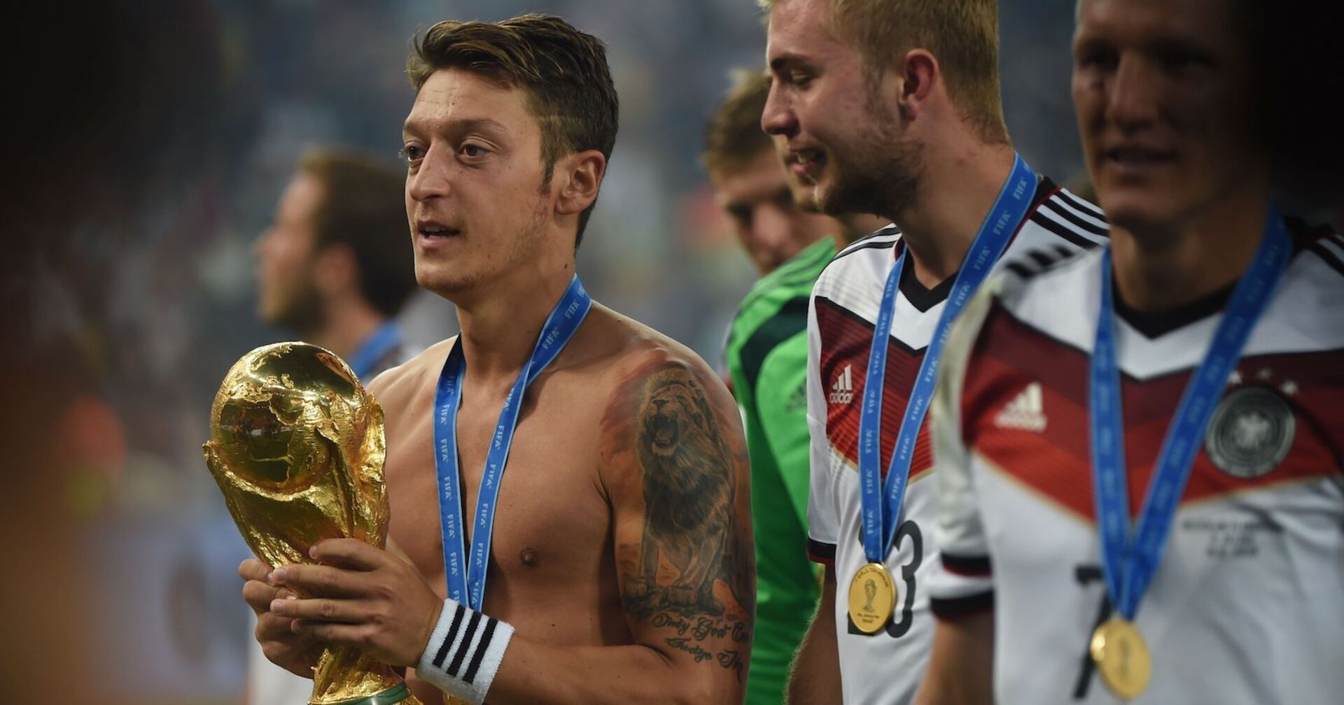 Mesut Özil hält die Weltmeisterkugel. Er hat kein Shirt an, auf seinem Arm ein großes Tattoo. Um den Hals die Medaille.