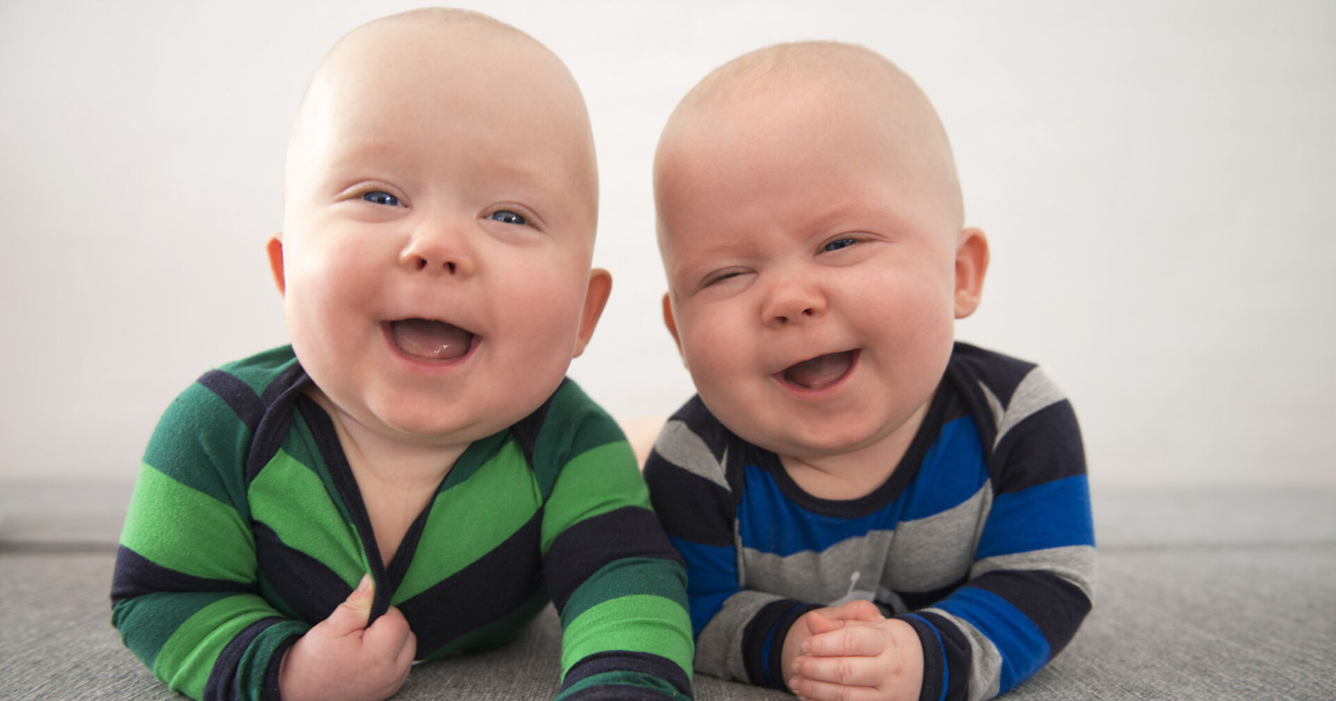 Zwei Babys liegen am Bauch und lachen in die Kamera.