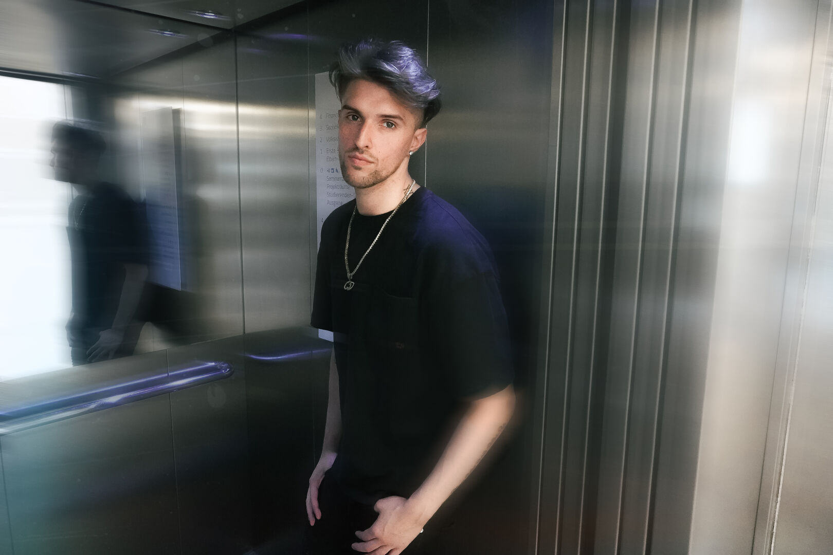 Gavis Dean, der - ganz in schwarz gekleidet und eine Goldkette um den Hals tragend -, in der Kabine eines Aufzuges steht