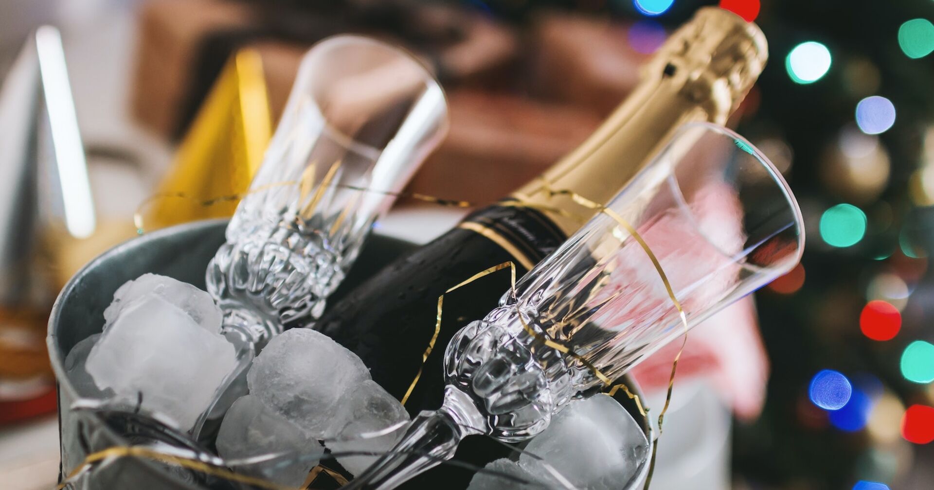 Eine geschlossene Sektflasche mit Sektkappe in einem Weinkühler mit Eis und zwei Gläsern.