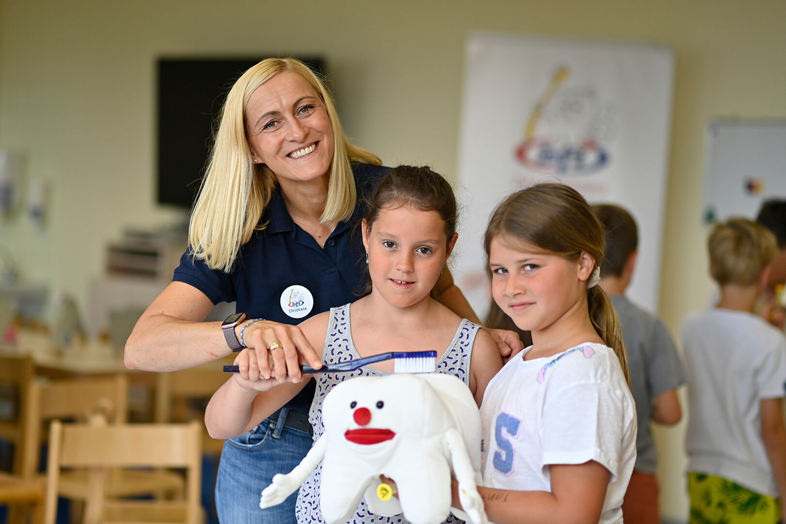 Eine Frau zeigt zwei Mädchen, wie sie richtig Zähne putzen anhand eines Maskottchens (Backenzahn).