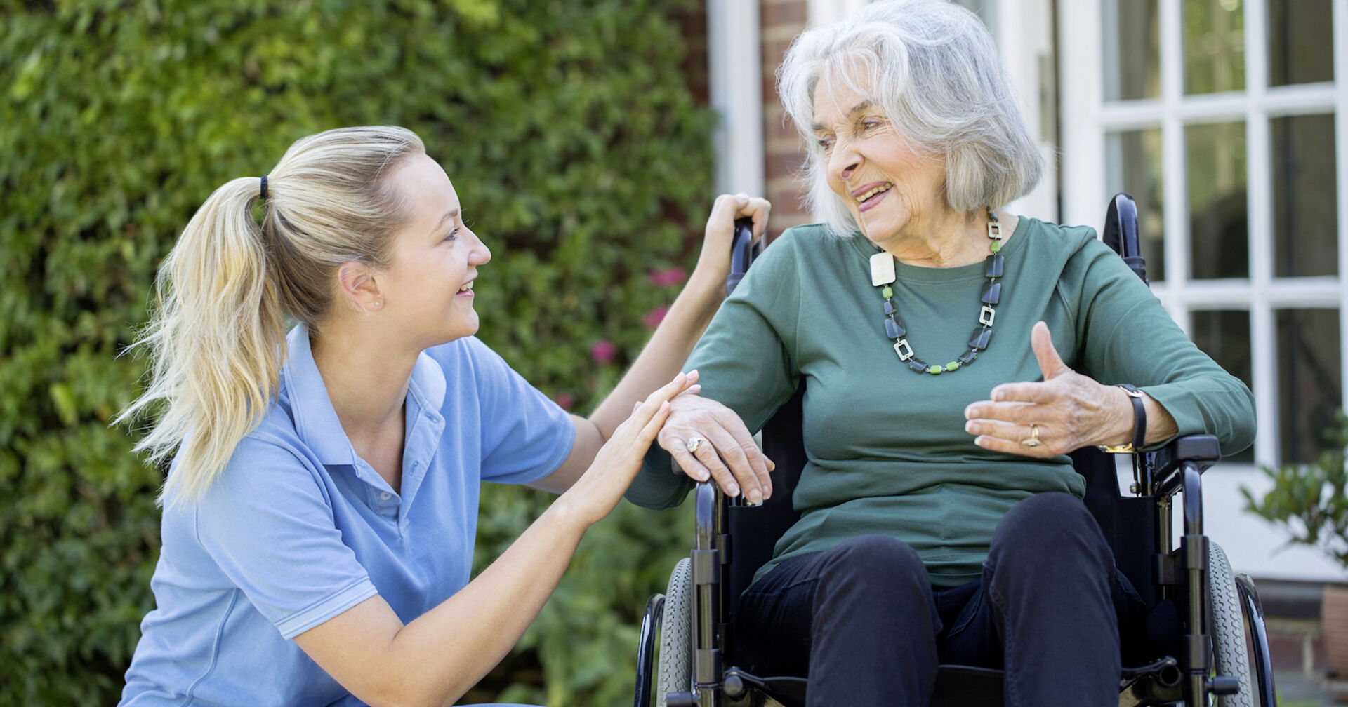 Junge Pflegerin kniet neben älterer Dame, die im Rollstuhl sitzt