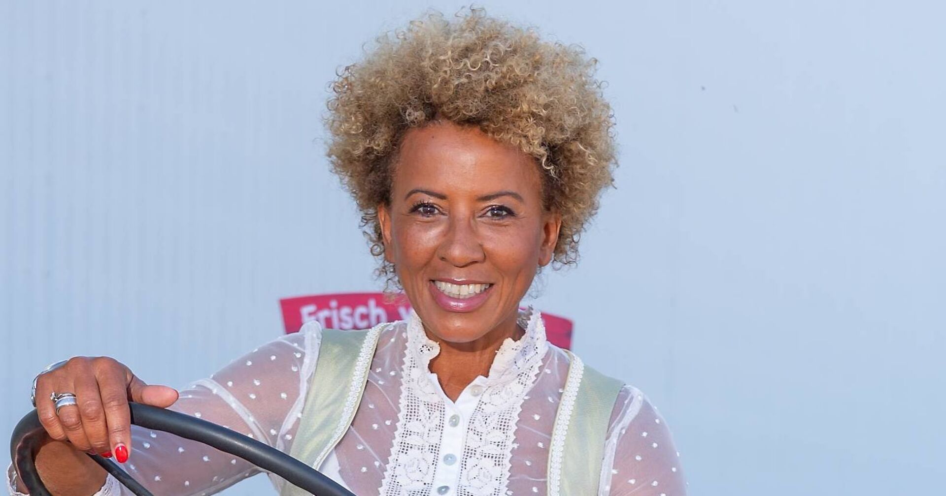 Arabella Kiesbauer in einem Dirndl mit blond gefärbten Haaren vor einer weißen Wand