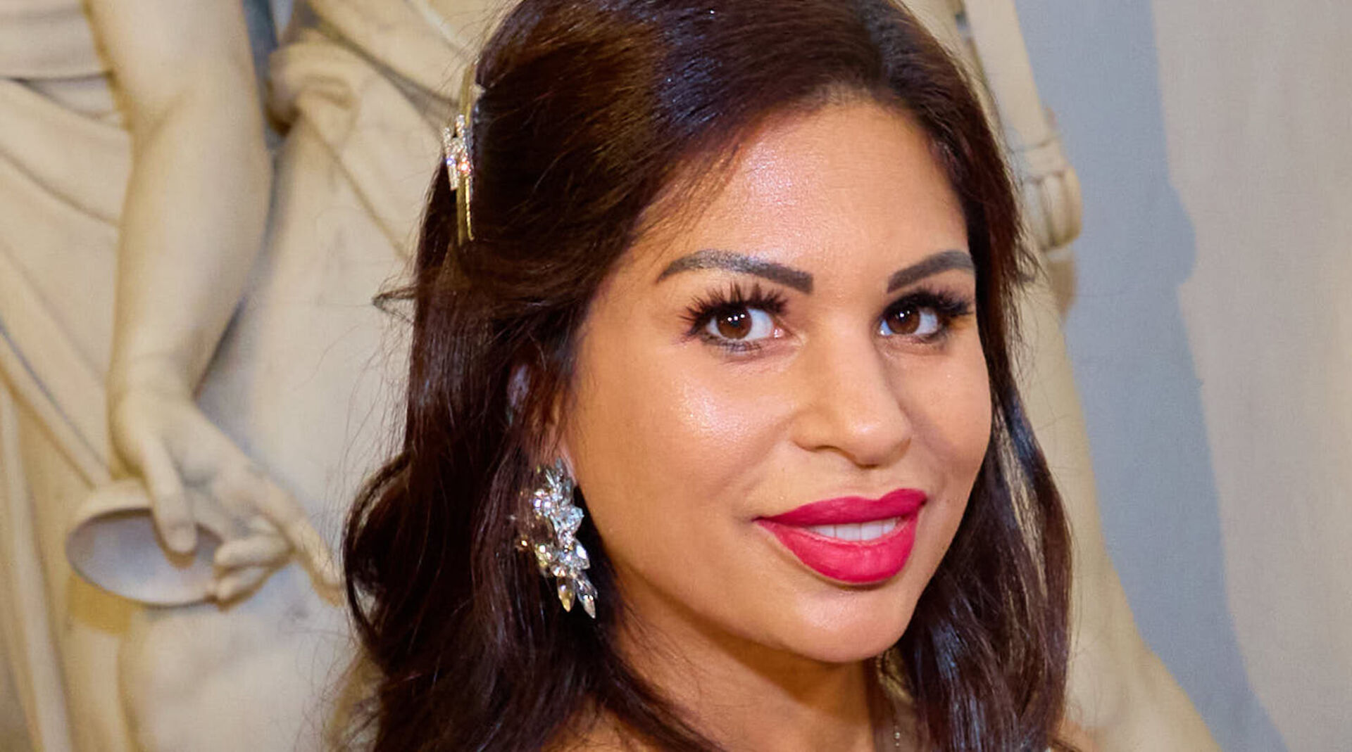 Nina Bambi Bruckner vor einer weißen Statue mit pinken Lippenstich und großen Ohrringen.