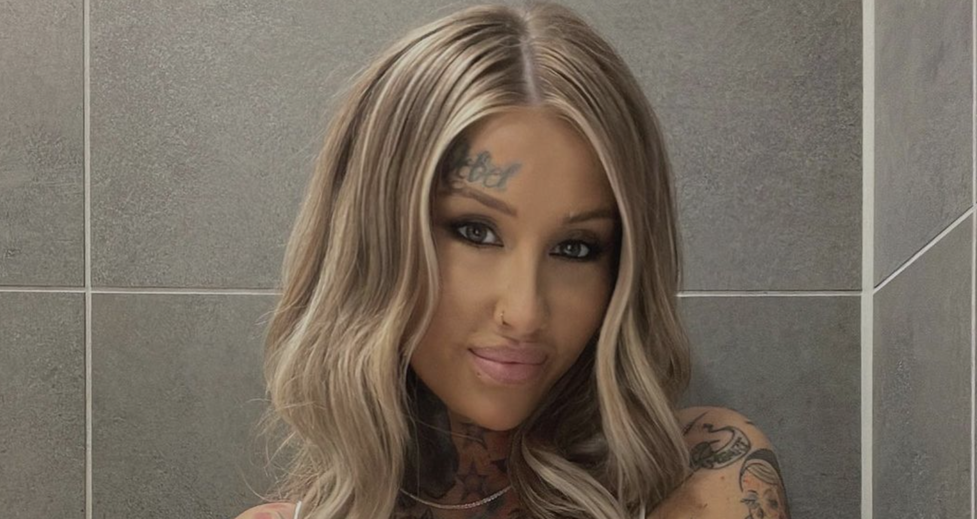 Blonde Frau mit Tattoo im Gesicht vor einer Fliesenwand