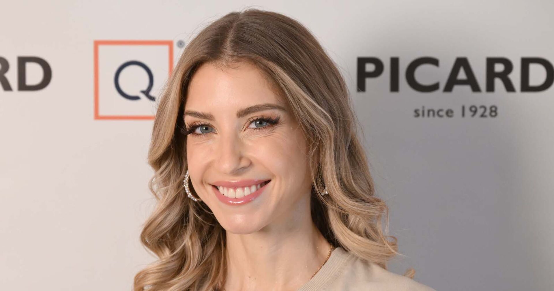 Cathy Hummels mit offenen blonden Haaren vor einer Fotoleinwand bei einem Event.