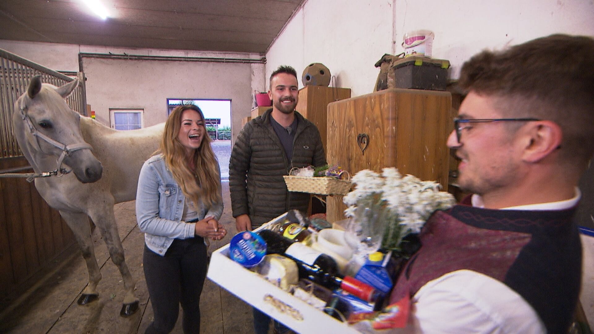 Ein Pferd steht hinter einer Frau mit langen blonden Haaren. Sie lacht und wird gerade von einem Mann reichlich beschenkt. Hinter ihr steht auch ein Mann und schaut zu.