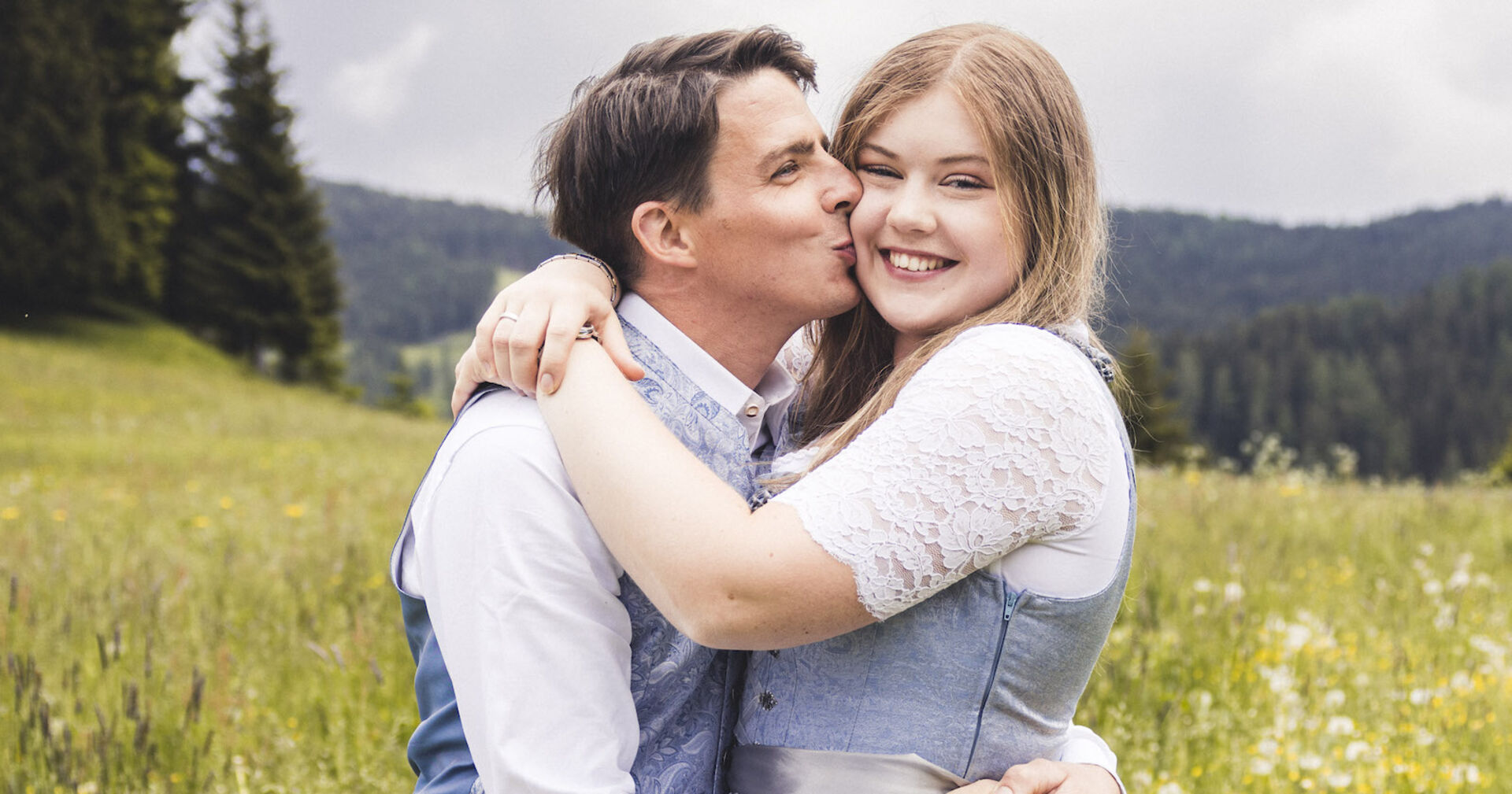 Ein Mann in Tracht gibt einer Frau im Dirndl mit blonden Haaren einen Kuss auf die Wange. Sie stehen auf einer Wiese.
