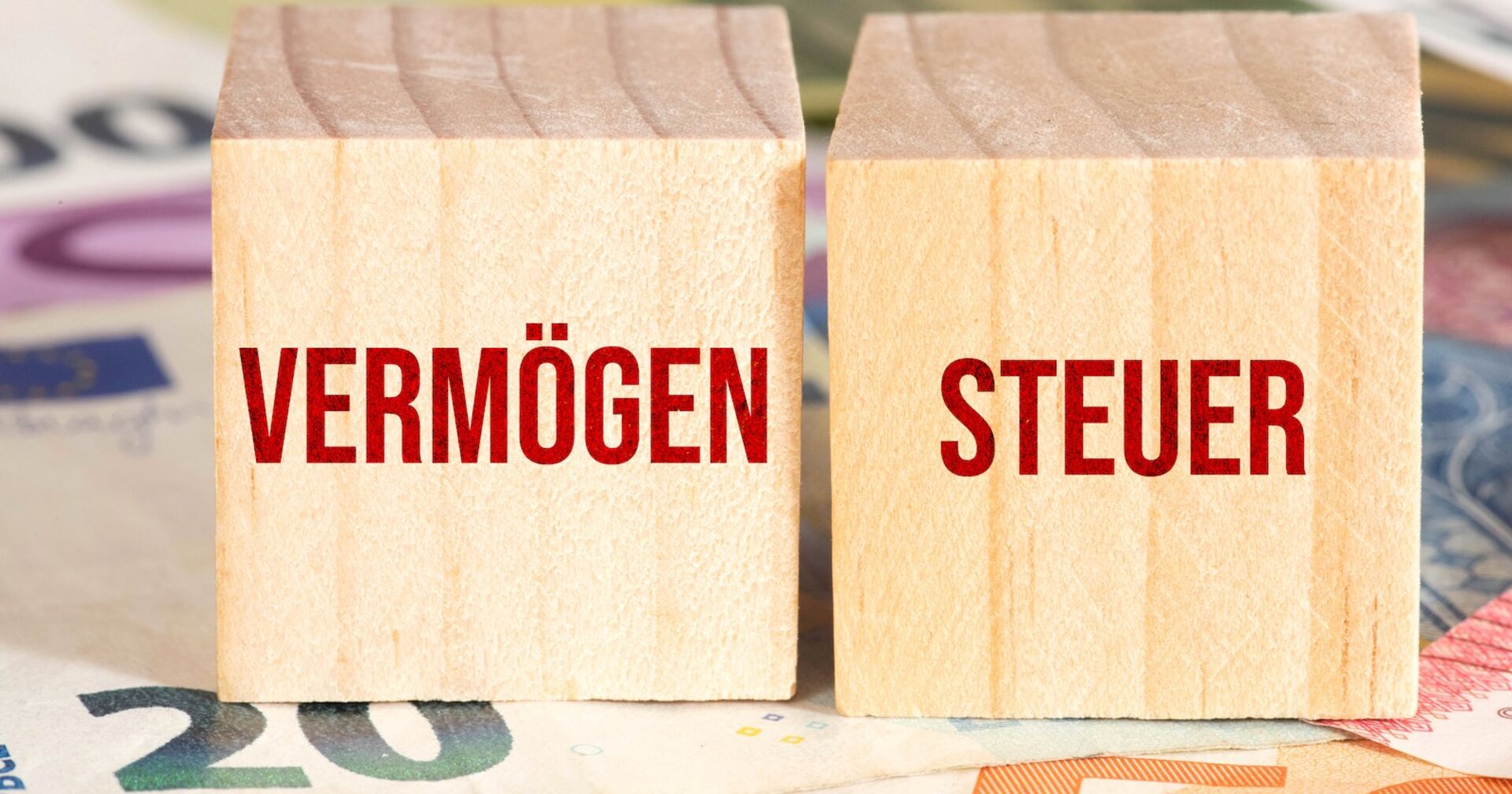 Zwei Holzwürfel, die auf mehreren Euro-Geldscheinen stehen und mit den Wörten Vermögen und Steuer versehen sind