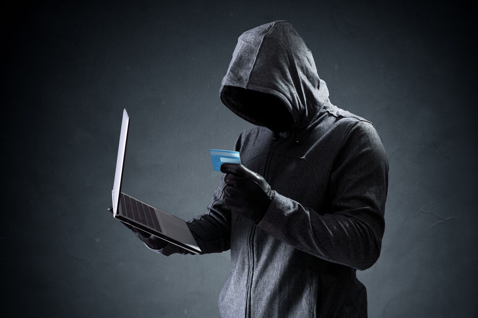 Ein Hacker in einem schwarzen Kapuzenpullover, der einen Laptop und eine Kreditkarte in Händen hält