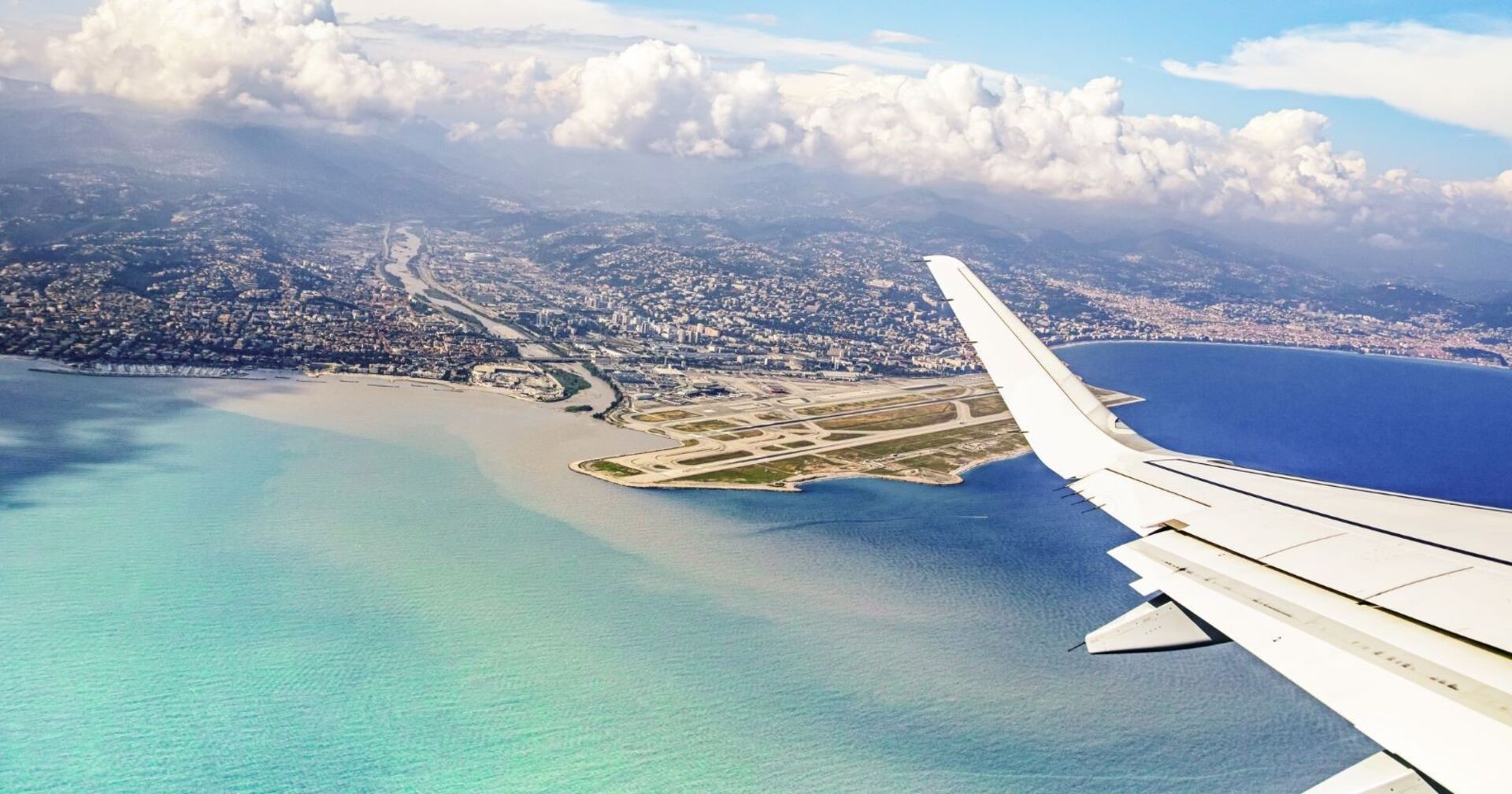 Die Tragfläche eines Flugzeugs aus dem fliegenden Flugzeug fotografiert. Darunter türkises Meer und eine Landebahn